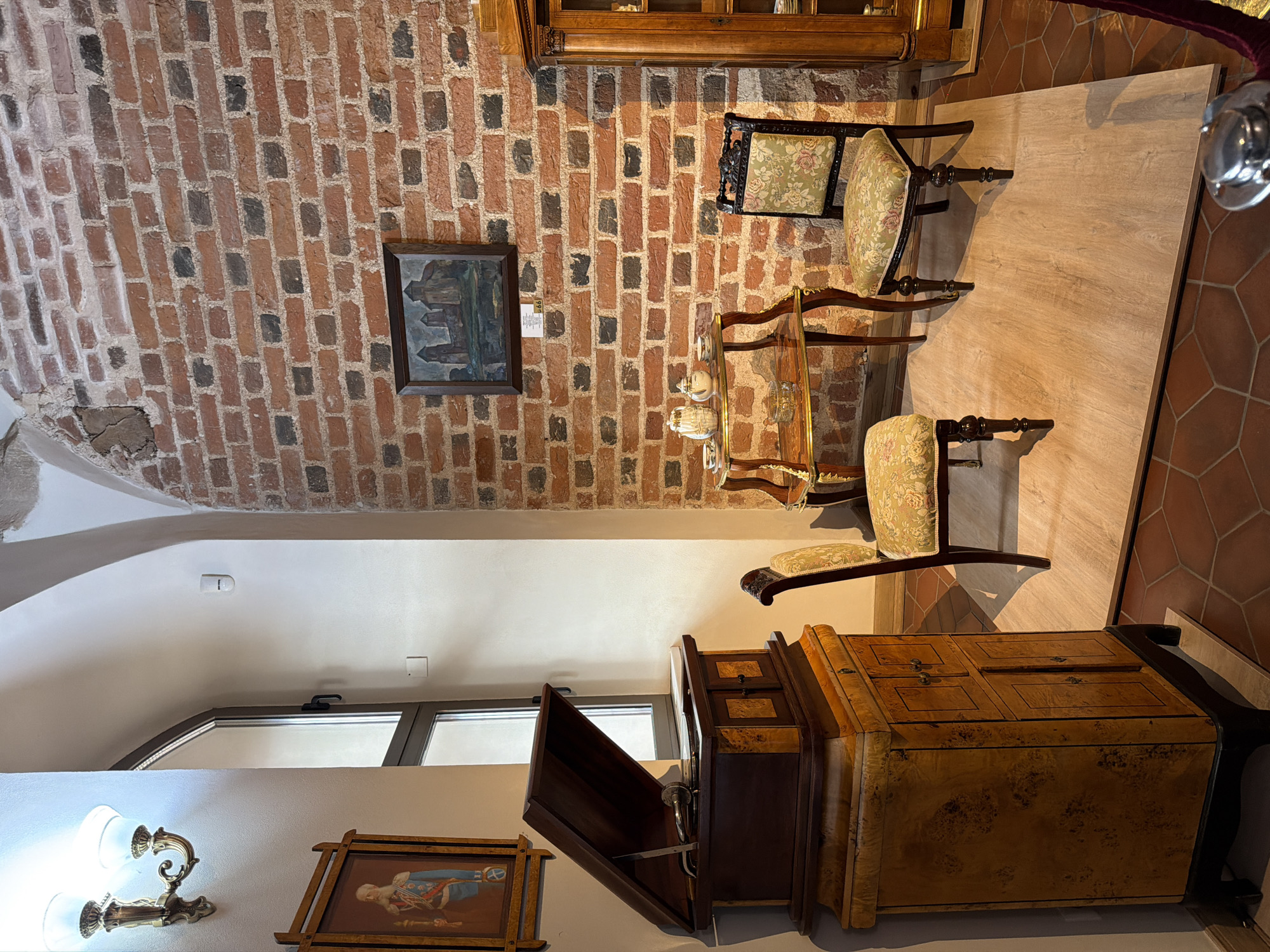 The image depicts an indoor scene, likely a museum or antique shop, showcasing a collection of vintage furniture and decor. A prominent feature is the exposed brick wall behind several pieces. On the wall, a small framed painting hangs, depicting buildings.
Several antique chairs with floral upholstery are arranged around a small, ornate glass-top table. A larger, ornate wooden cabinet with a lift-top is positioned in the foreground. Beside it, another antique cabinet, perhaps a secretary desk, is visible with its drawers partially open, revealing an interior with a mechanism.
To the left, a wall-mounted light fixture with two glass shades is visible, along with a framed painting of a person in a historical military uniform. The floor has a tiled pattern. The lighting suggests it is daytime, with light coming from windows that are partially visible and also from overhead fixtures. The scene conveys a sense of history and an appreciation for antique craftsmanship. No discernible text is visible.