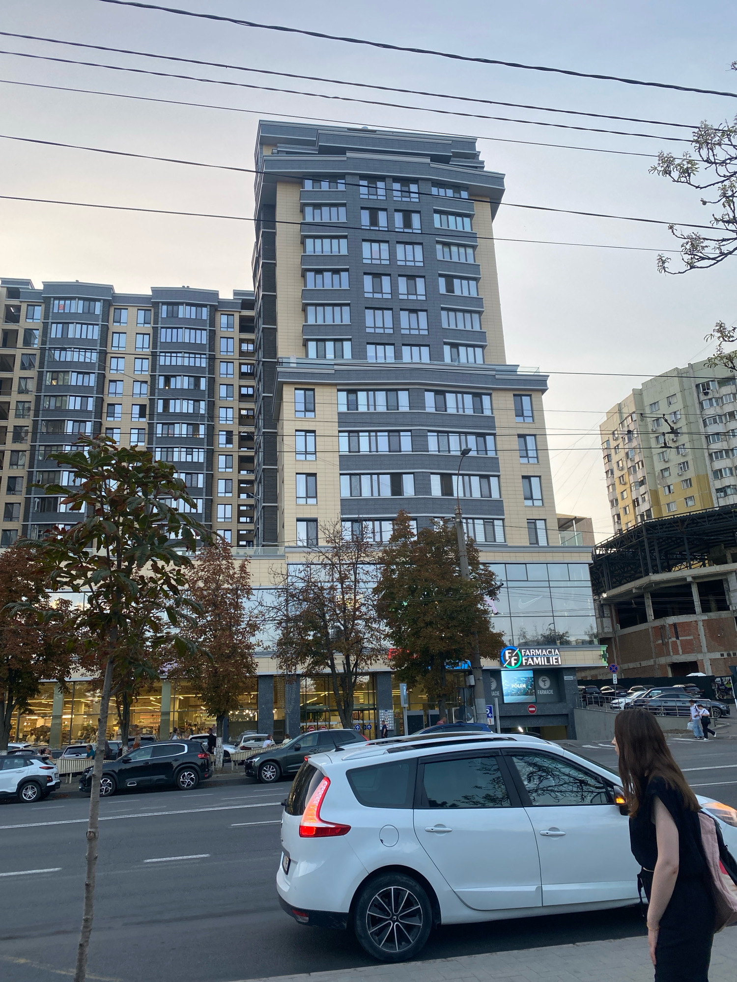 The image captures a street scene in Chișinău, Moldova, with a prominent modern apartment building as the focal point. The building features a mix of grey and beige facade elements with numerous windows. In the foreground, a white station wagon is parked on the right side of the street, partially obscuring the view of a woman in a black top and skirt who is facing away from the camera. Other vehicles, including a black SUV and several other cars, are visible parked along the street and in front of a large retail establishment with illuminated windows. Trees with autumnal foliage line the sidewalk. Overhead, power lines crisscross the sky, which is overcast. To the right of the main apartment building, another, older apartment building is visible, as well as a construction site with scaffolding. A sign for "FARMACIA FAMILIEI" (Family Pharmacy) is displayed prominently on a digital billboard. The overall impression is of an urban environment during daytime.