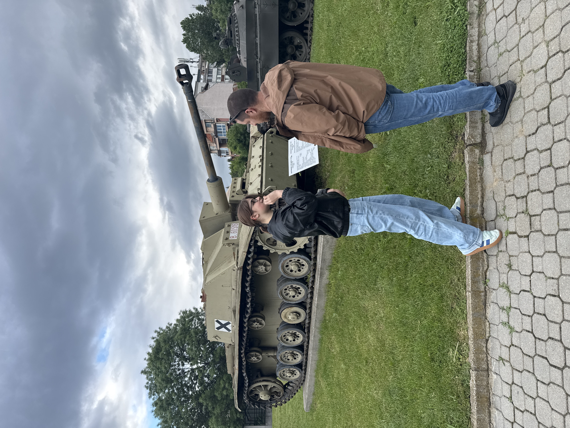 Two individuals, a man and a woman, are standing near a military vehicle in an outdoor setting. The man wears a brown jacket, blue jeans, and black shoes. He is peering at a sign near the vehicle. The woman is dressed in a black jacket and blue jeans, and is looking upwards at the military vehicle. The military vehicle, which appears to be a self-propelled gun or tank destroyer, is positioned on a grassy area next to a paved walkway. A second military vehicle is visible in the background. The setting is likely a museum or memorial site. The location is Sofia, Bulgaria.