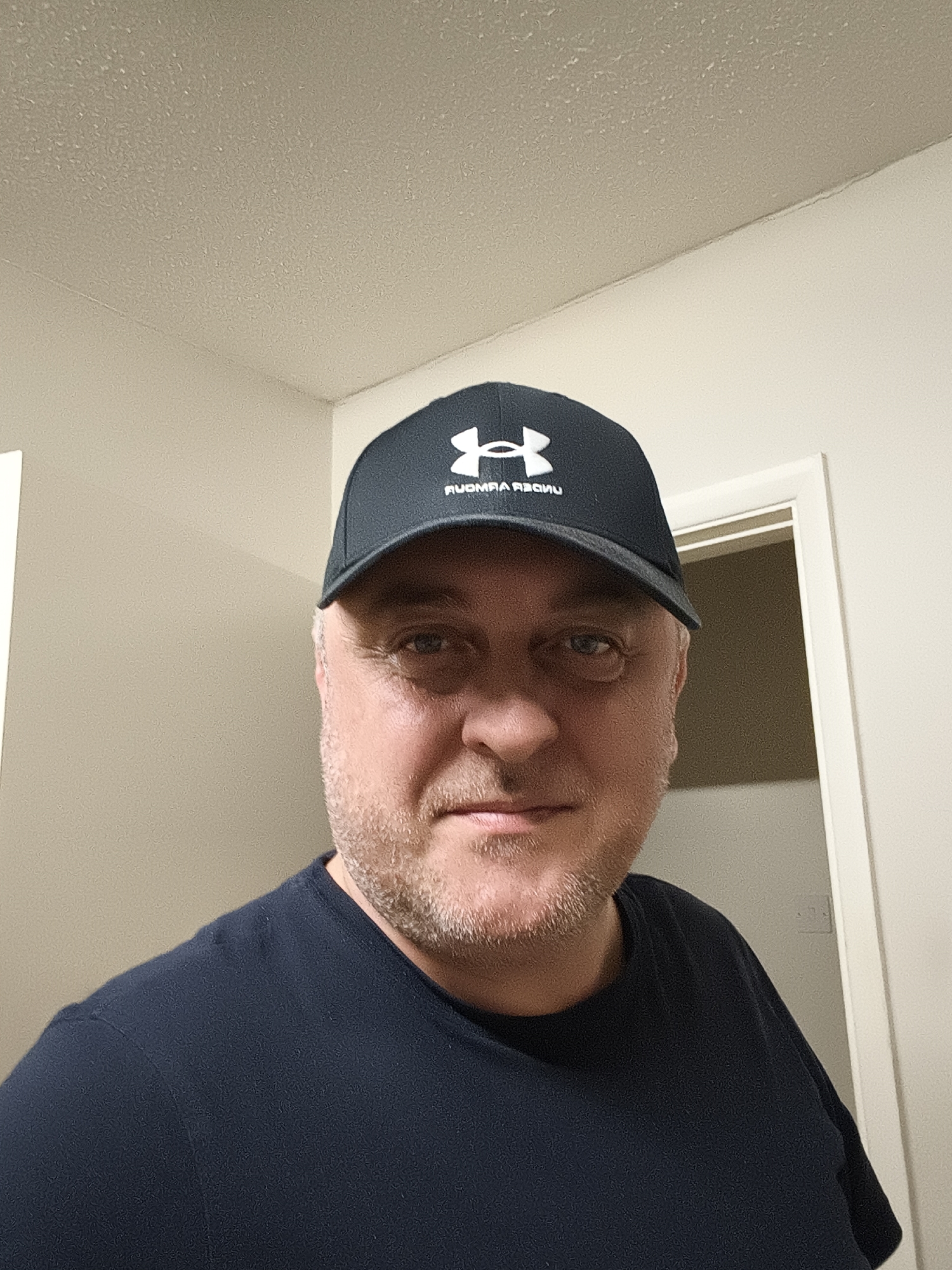 A close-up, eye-level portrait shows a middle-aged man indoors. He is looking directly at the camera with a neutral to slightly content expression. The man has fair skin, light-colored stubble on his face, and visible bags under his eyes. He is wearing a dark blue or black crew-neck t-shirt and a dark baseball cap. The cap features a white "H" shape logo and the mirrored text "RUOMRA REDNU", indicating it is an "Under Armour" cap, likely captured with a front-facing camera.
The background consists of light-colored walls, possibly off-white or light beige, and a textured, potentially popcorn-style ceiling. To the man's right (viewer's left), a corner of a wall and ceiling is visible. To his left (viewer's right), there is a door frame, with the interior of the adjacent room appearing darker. The lighting is artificial, illuminating the scene brightly. The setting appears to be a residential or commercial indoor space in Huntingdonshire, United Kingdom, captured in what seems to be a selfie.