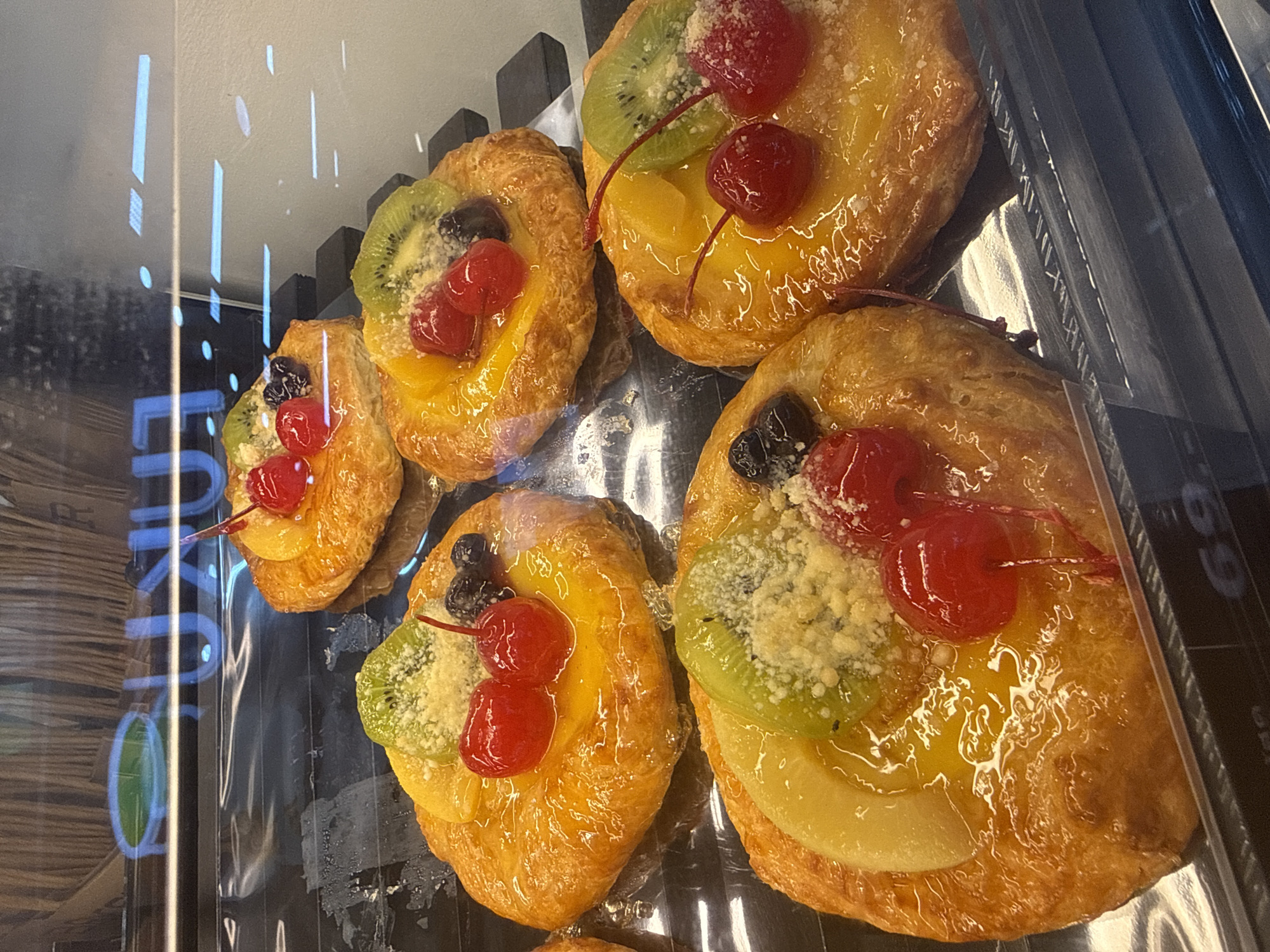 In Pattaya City, Thailand, a close-up, eye-level shot showcases an assortment of fruit-topped pastries displayed in a glass-fronted refrigerated cabinet, likely in a bakery or market. The pastries, approximately five visible, are round and golden-brown with a flaky crust, generously topped with a glossy yellow glaze. Each pastry features a garnish of sliced kiwi fruit, a few dark blueberries, and two bright red cherries with stems. Some pastries also have a sprinkle of grated cheese or a similar white crumbly topping. The lighting highlights the sheen of the glaze and the textures of the fruits and pastry. Reflections of the store's interior, including blue lettering and overhead lights, are visible on the glass surface. The price tag "69" is partially visible at the bottom right corner.