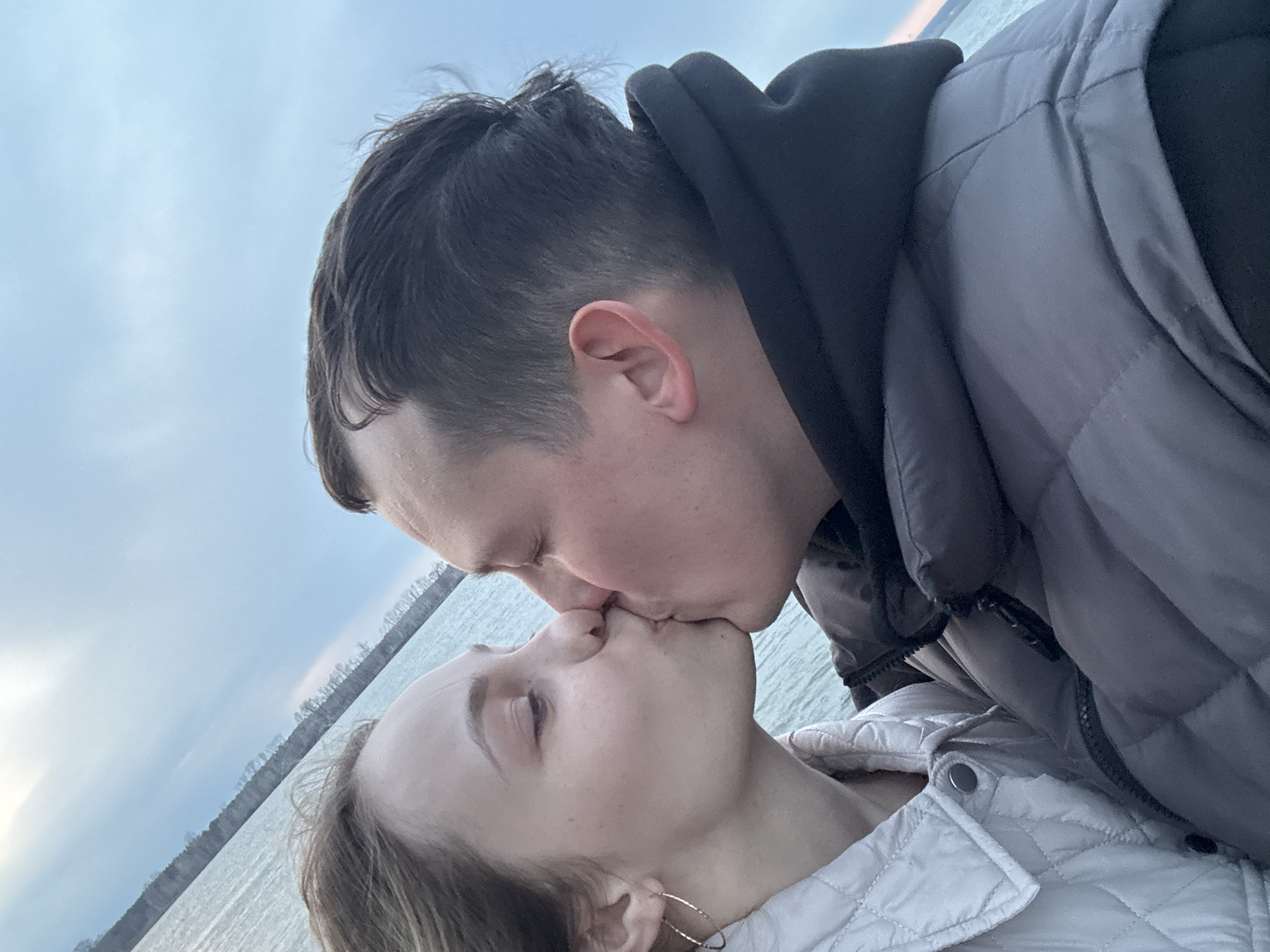 This image captures a close-up, tilted shot of a couple sharing a kiss outdoors. The man is positioned in the upper right portion of the frame, leaning down towards the woman who is in the lower left, looking up. He has dark hair and fair skin, and is wearing a dark hooded sweatshirt underneath a grey padded jacket. The woman has lighter hair and fair skin, and is dressed in a light-colored, possibly white or light grey, quilted jacket, with a gold hoop earring visible on her right ear. Both individuals have their eyes closed, indicating a moment of intimacy and affection.
The background shows a large body of water, likely a lake or wide river, with a distant tree line visible along the far shore. This setting is consistent with the location context of Chaikovsky, Russia, which is situated near the Kama River and Votkinsk Reservoir. The sky is overcast or partly cloudy, providing soft, diffused lighting. The time of day appears to be daytime, possibly late afternoon or early evening, given the gentle light. The overall mood is romantic and tender. No visible text is present in the image.