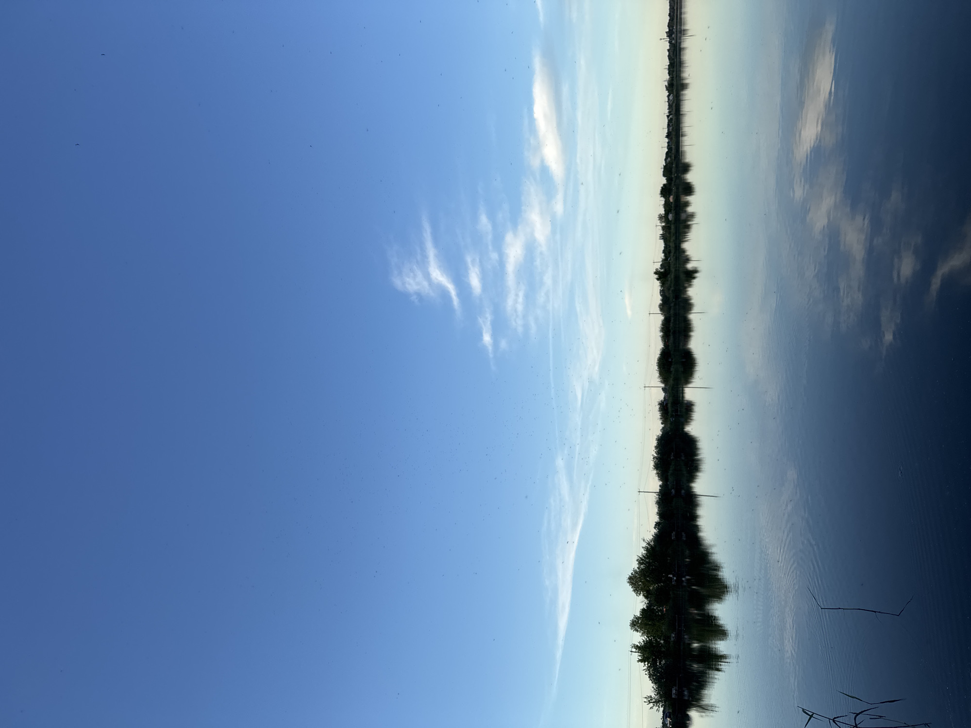 The image depicts a body of water reflecting a shoreline of trees and a cloudy sky. The water surface appears calm, mirroring the trees and clouds above. The sky is predominantly blue with some white clouds. The shoreline consists of trees and possibly some buildings, though the details are somewhat obscured by the reflection. The location appears to be near water.