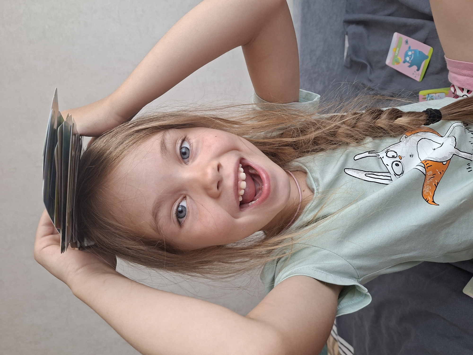 A young girl with blonde hair styled in a braid is sitting indoors, holding a stack of cards on top of her head. She is wearing a light green t-shirt with a rabbit graphic and pink shorts. Her mouth is open in a wide smile, revealing her teeth. In the background, a gray sofa and a wall are visible, along with a couple of small cards on the sofa. The image appears to be taken in a domestic setting.