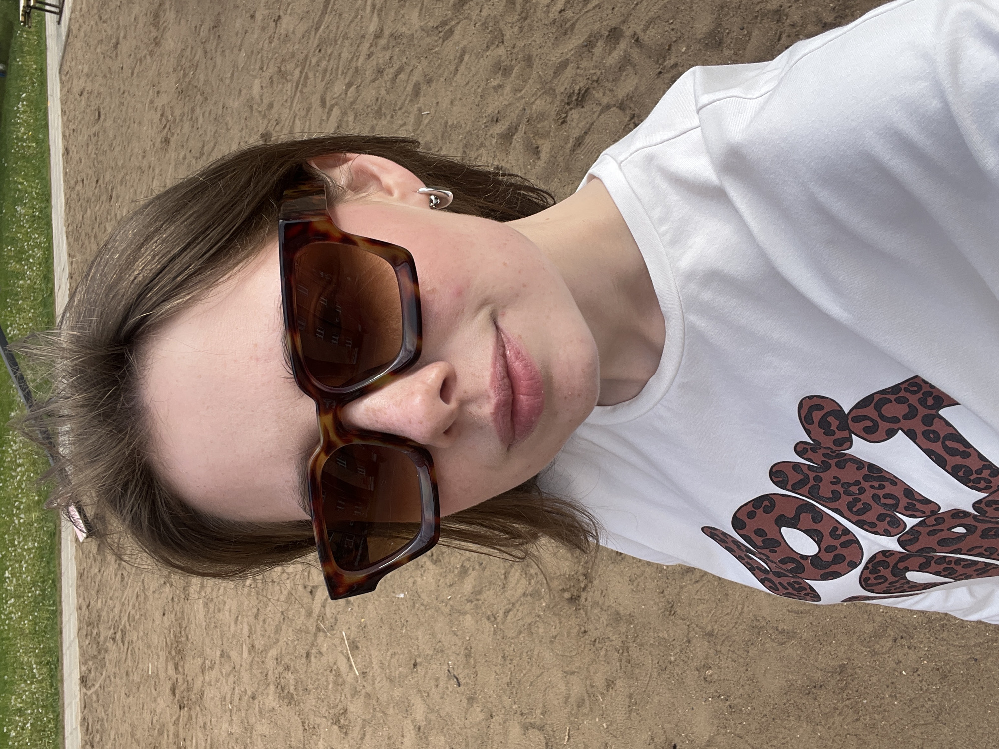 A close-up, angled portrait captures a young person lying on sandy ground in Chaikovsky, Russia, during a sunny daytime. The image is oriented counter-clockwise by 90 degrees. The person has light brown hair and is wearing oversized, square-framed tortoiseshell sunglasses, which obscure their eyes. A small silver-toned earring is visible on their right ear. Their lips are slightly parted, displaying a neutral expression. They are dressed in a white short-sleeved t-shirt featuring a distinctive graphic: large, brown letters with black leopard spots that clearly spell "NO IDEA."
The background primarily consists of light brown, dry, sandy earth, suggestive of a beach, playground, or open outdoor area. To the left of the frame, a strip of vibrant green grass speckled with small white flowers is visible. Reflections in the sunglasses show what appears to be a building with multiple windows, hinting at an urban or developed setting nearby. The bright sunlight creates clear lighting conditions, emphasizing the textures of the sand and the details of the subject's attire.