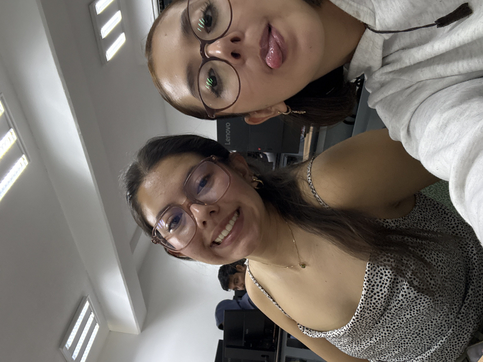 This image is a selfie featuring two young women, taken from a slightly upward angle in an indoor setting. The woman in the foreground is smiling warmly, looking directly at the camera. She wears glasses with light brown frames, a delicate gold chain necklace with a small green pendant, and a white and black spotted strap dress. Her long, dark hair falls over her left shoulder.
Behind her, and positioned higher in the frame, another woman is also wearing glasses with darker frames. She is playfully sticking her tongue out towards the camera. She wears a light-colored, possibly grey, hoodie. Both women have dark hair and appear to be in their late teens or early twenties.
In the background, a man is partially visible sitting at a desk, facing away from the camera. The setting appears to be a classroom, office, or computer lab, characterized by bright fluorescent strip lights on the ceiling and computer monitors. One monitor clearly displays the "Lenovo" brand logo. The overall atmosphere conveyed is casual and lighthearted. The location of this scene is San Andrés Cholula, Mexico.
