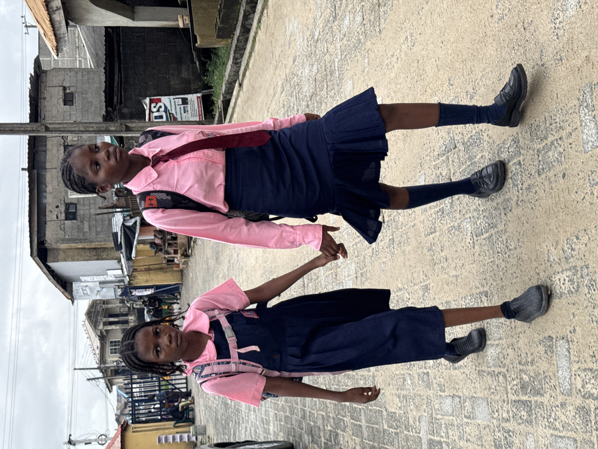 The image, which is rotated 90 degrees clockwise, depicts two young girls in school uniforms standing side-by-side on a textured paved surface. Both girls have dark skin and braided hair.
The girl on the left wears a pink long-sleeved shirt, a burgundy tie, a navy blue pleated skirt, dark socks, and black lace-up shoes. She has a dark backpack with pink and white accents on her back. Her expression is neutral as she looks slightly off-camera.
The girl on the right is also dressed in a pink long-sleeved shirt, worn under a navy blue sleeveless overall dress with pink straps. She wears dark socks and gray slip-on shoes, and carries a dark backpack with a patterned section. Her gaze is directed towards the camera with a neutral expression. The two girls are holding hands between them.
The background reveals a street scene with concrete block buildings, some painted and others unpainted, typical of an urban or semi-urban area in Ado, Nigeria. Utility poles and overhead wires are visible. The sky is overcast, indicating daytime. On a building behind the girl on the left, a white sign with red and black text is partially visible, showing characters like "OS" and part of a phone number starting with "090". The scene suggests the girls are on their way to or from school.