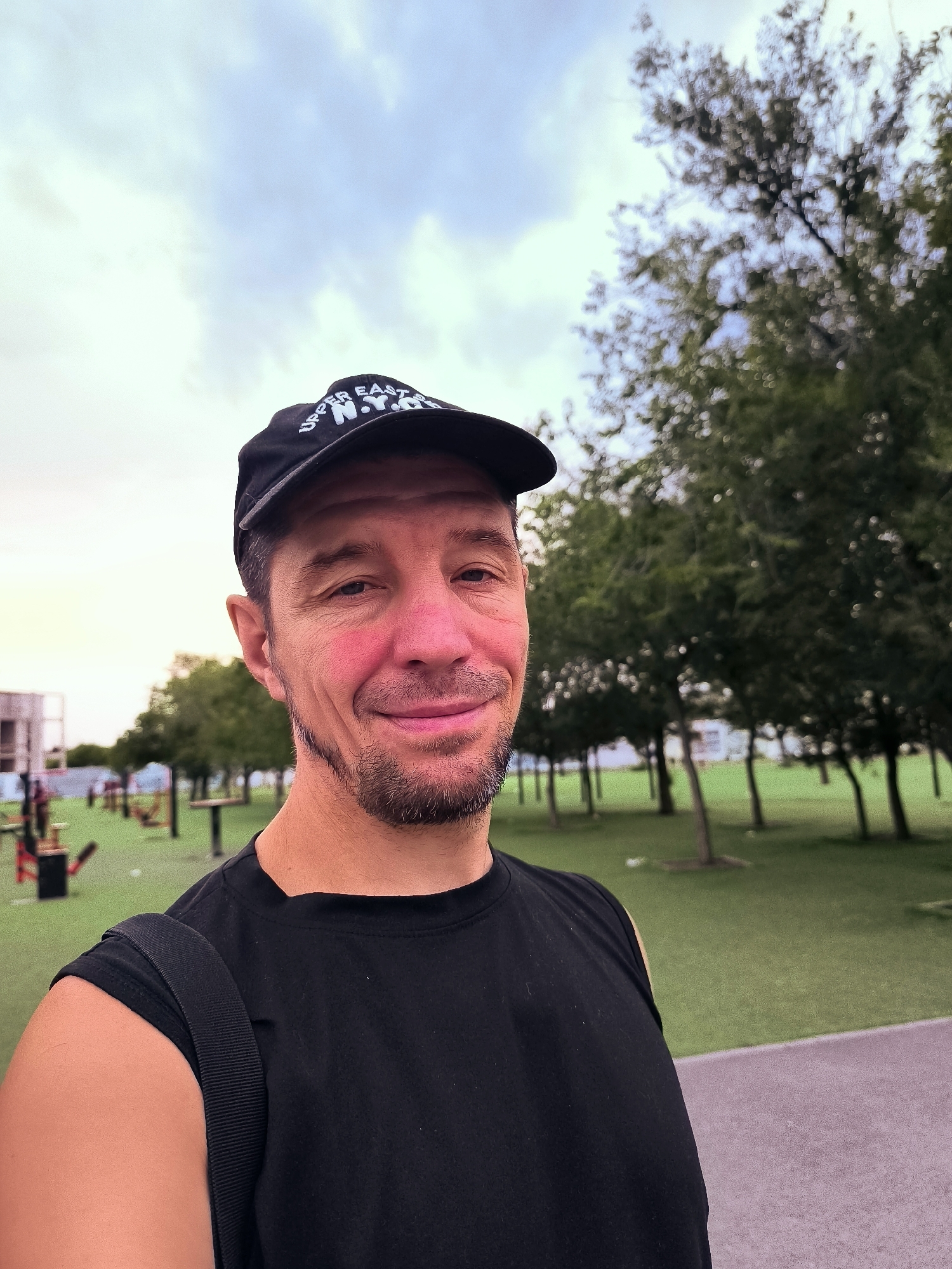 A man in a black t-shirt and a black baseball cap with "UPPER EAST N.Y.C." written on it is taking a selfie outdoors. He has short, dark hair and a goatee. His face is slightly flushed. The background shows a park with trees and exercise equipment. The sky is cloudy.