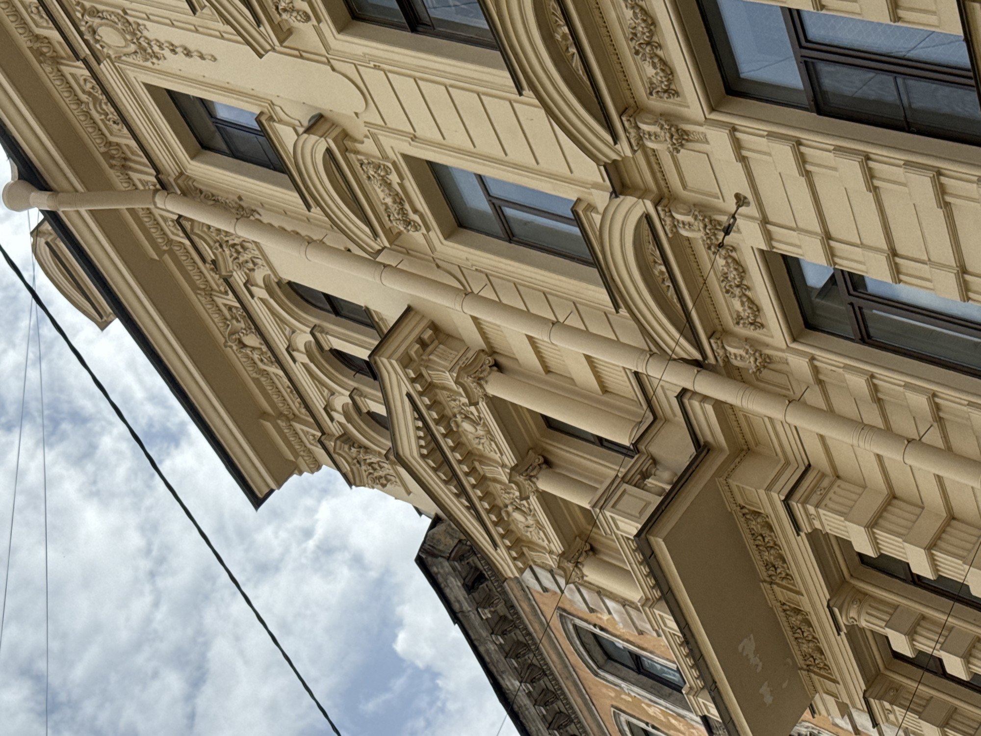 The image presents a low-angle view of the facade of a building, likely located in Saint Petersburg, Russia, based on the architectural style. The building is characterized by its ornate decorations, featuring detailed moldings, window frames, and sculptural elements. The primary color of the facade is a creamy, light yellow. Windows are rectangular and reflect the sky. A rain gutter runs along the edge of the building. A cloudy sky is visible in the lower left corner of the image, which is otherwise focused on the architectural details.