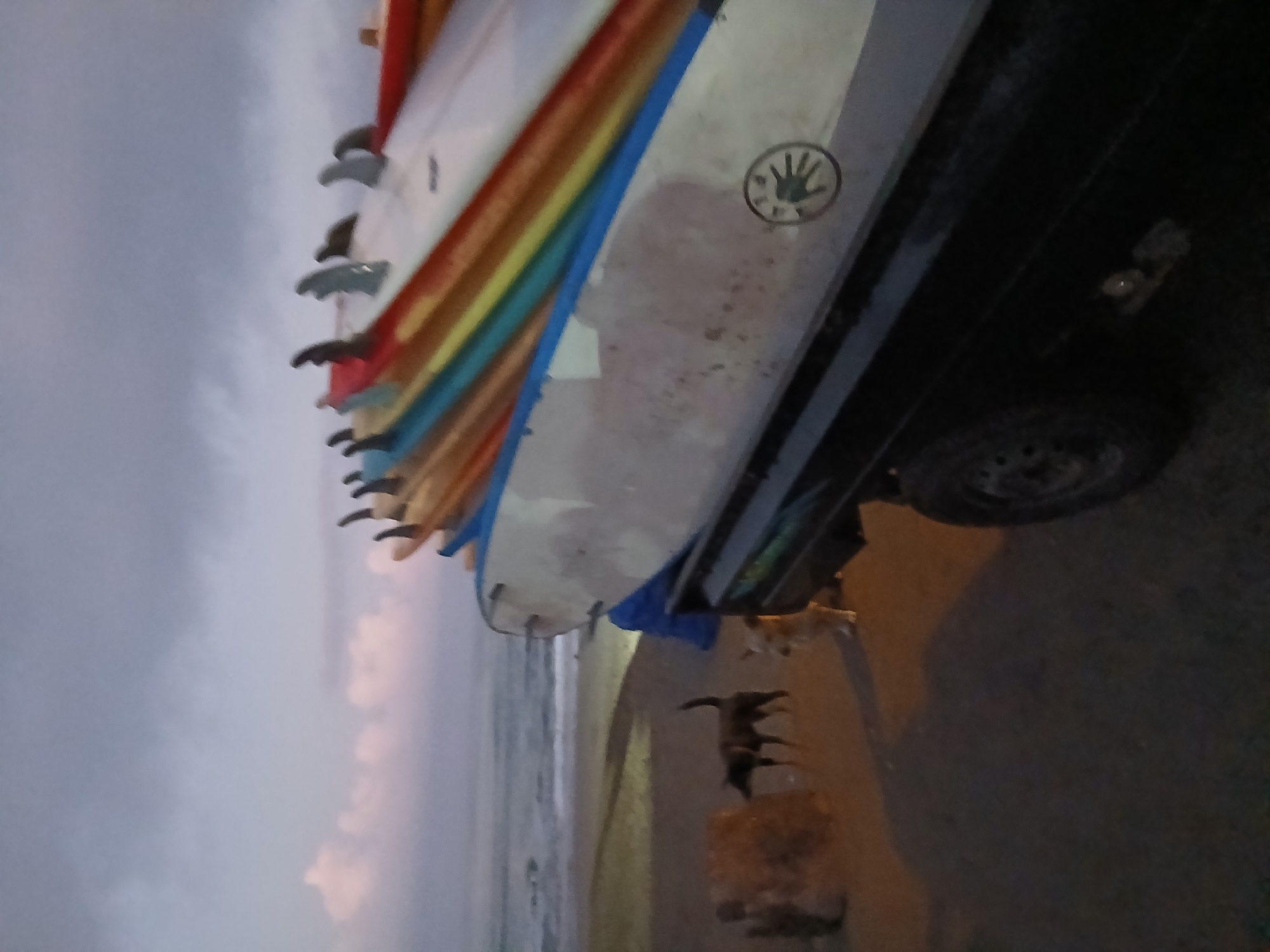 At dusk on a beach in North Kuta, Indonesia, a flatbed truck is parked on the sand. The truck is laden with a stack of surfboards of varying colors, including blue, yellow, red, and white. The surfboards are angled upwards and to the right. Two dogs are present on the beach; one is dark-colored and standing, while the other is lighter and sitting. The ocean is visible in the background, with gentle waves lapping at the shore. The sky is overcast with hints of pink and orange from the setting sun. The overall scene suggests a surf-related activity or transport of surfboards at the end of the day.