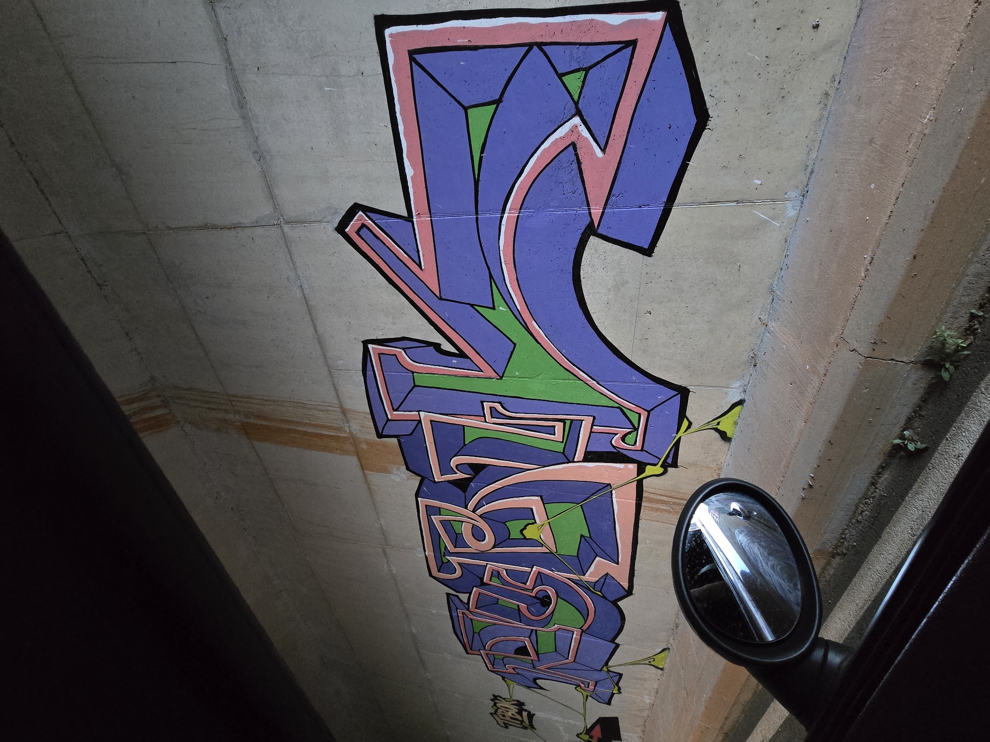 The image depicts a concrete wall, likely part of an underpass or bridge in the city of Klopot, Montenegro, adorned with vibrant graffiti. The graffiti artwork features stylized letters in shades of purple, pink, and green, with bold black outlines and three-dimensional shading. The style suggests a "wildstyle" or complex bubble letter graffiti, common in urban art. The wall itself shows signs of age and weathering, with streaks of orange discoloration running from the top. To the left of the graffiti, a small tag that reads "TPOK" is visible. In the foreground, a car's side mirror is visible, reflecting a dimly lit road and the car's interior. The overall environment appears to be a somewhat neglected urban area, possibly a public space or transportation infrastructure.