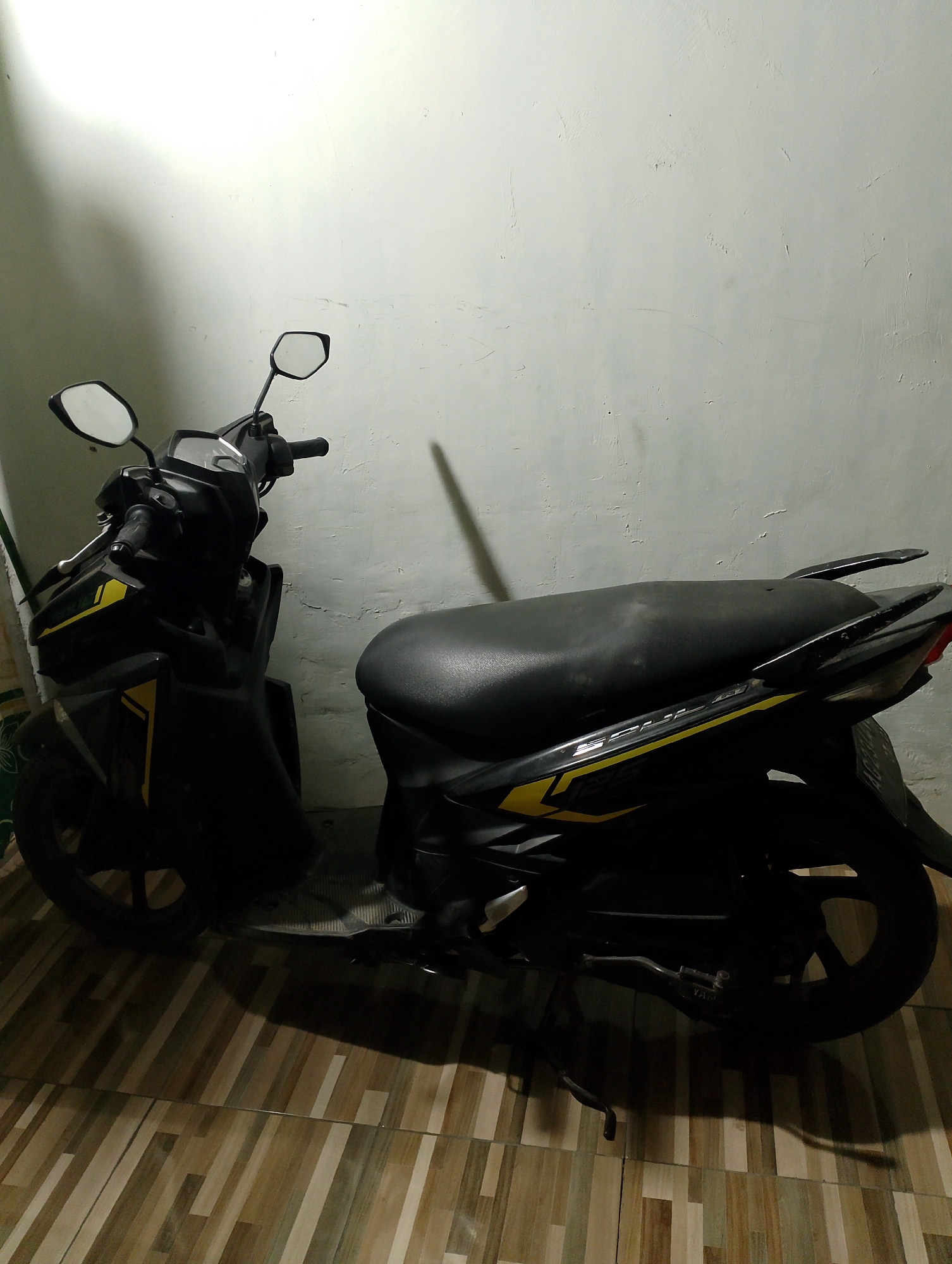 A black Yamaha Mio scooter is parked indoors against a lightly textured, off-white wall. The scooter has yellow decals with the word "SOUL" visible on its side. The floor is covered with rectangular tiles in varying shades of brown and beige, laid in a staggered pattern. The lighting casts a strong shadow of the scooter on the wall. The scene appears to be in a residential setting.