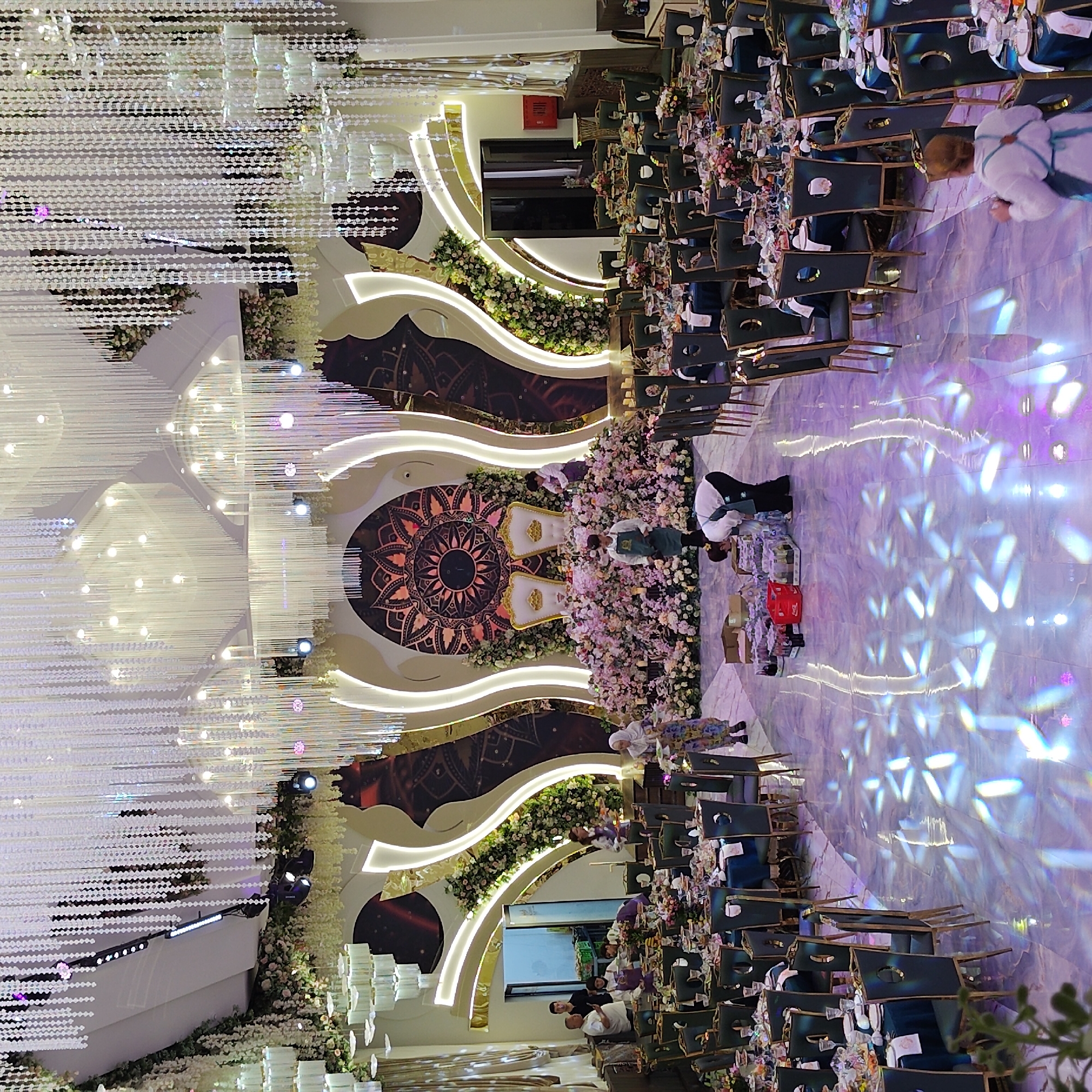 This image depicts the interior of a lavishly decorated banquet hall, likely in Khujand, Tajikistan, set up for a celebratory event, possibly a wedding. The hall features an abundance of floral arrangements, particularly a massive display of pink and white flowers surrounding a decorated backdrop with two ornate chairs at the center, suggesting a ceremonial seating area.
The ceiling is adorned with numerous crystal chandeliers and hanging decorative elements, creating a dazzling effect. The floor is a polished, reflective surface with scattered light projections, adding to the opulent ambiance.
Several round tables, draped in dark blue cloths and set with place settings and centerpieces, fill the foreground and midground, indicating that guests are either seated or about to be seated. Dark chairs with gold accents surround the tables.
In the midground, several individuals are visible. Some appear to be staff members attending to preparations. One person is seen handling drinks and boxes on the floor. Others are interacting with the tables or near the stage area. A woman in a colorful dress walks towards the right side of the image.
The overall impression is one of grandeur and meticulous preparation for a significant event. The backdrop features stylized, curved gold-trimmed panels and a large, circular mandala-like design. The lighting is a mix of ambient illumination from the chandeliers and spotlights, contributing to the festive and dramatic atmosphere. The presence of what appear to be food and drink preparations and staff suggests ongoing activity related to the event.