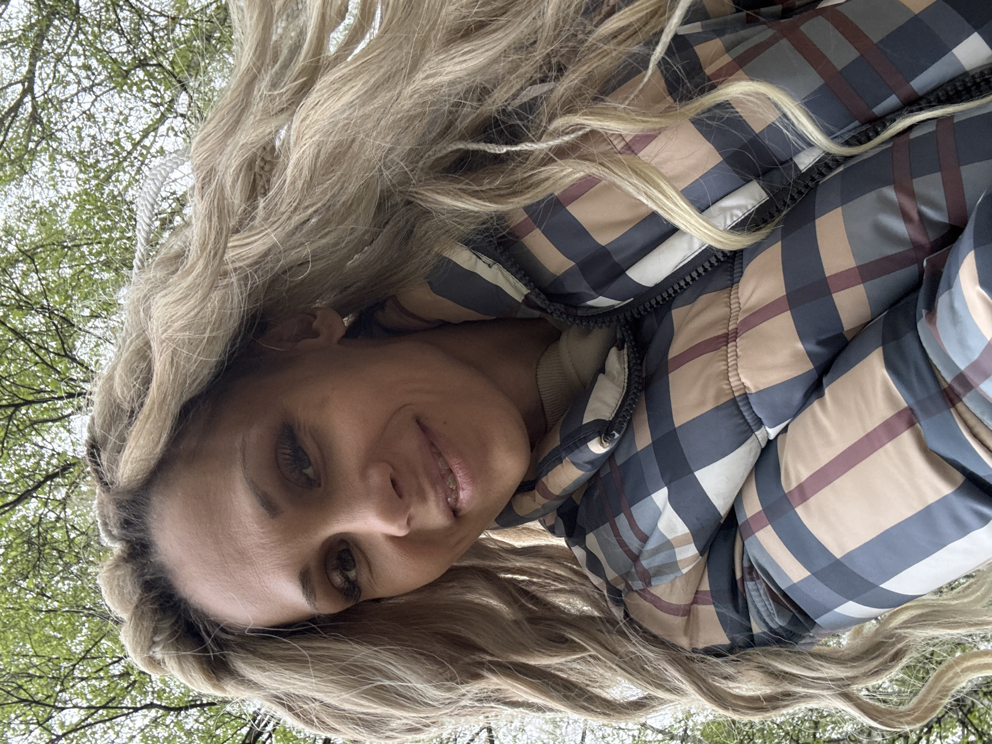 A woman is photographed from a low angle looking up. She has fair skin and long blonde hair, styled with braids and waves. She is wearing a jacket with a plaid pattern in shades of beige, blue, and maroon. The environment includes trees with green foliage, suggesting an outdoor setting. The photo appears to have been taken in a park or wooded area. There is no verifiable information about the location being in Rusanovo, Russia.
