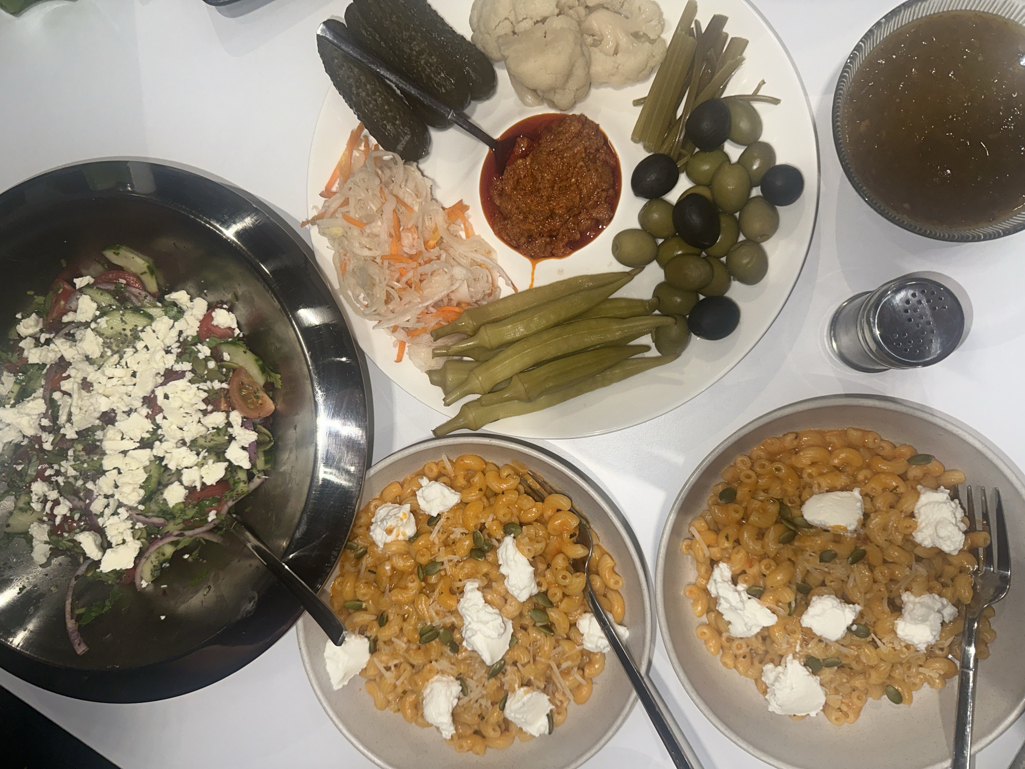An overhead view captures a dining table set with multiple dishes in Yerevan, Armenia. A large, polished metal bowl in the upper left section contains a salad with chopped cucumbers, red onions, tomatoes, herbs, and a generous topping of crumbled white cheese, with a serving spoon inserted. Adjacent to it, a white rectangular plate displays a variety of pickled and preserved items: shredded pickled cabbage and carrots, two whole pickled gherkins with a serving fork, a row of pickled okra pods, a central mound of red-orange paste or dip with a small spoon, pickled cauliflower florets, and a collection of green and black olives alongside thin pickled stems. In the lower right quadrant, a small glass bowl holds a brown, textured liquid, positioned next to a small metal shaker with a perforated top. Two identical beige ceramic bowls are present in the lower-left, each filled with orange elbow macaroni pasta, sprinkled with grated white cheese and green pumpkin seeds, and adorned with multiple dollops of white soft cheese. A metal fork rests in each pasta bowl. The table surface is uniformly white. No individuals are visible in the frame.