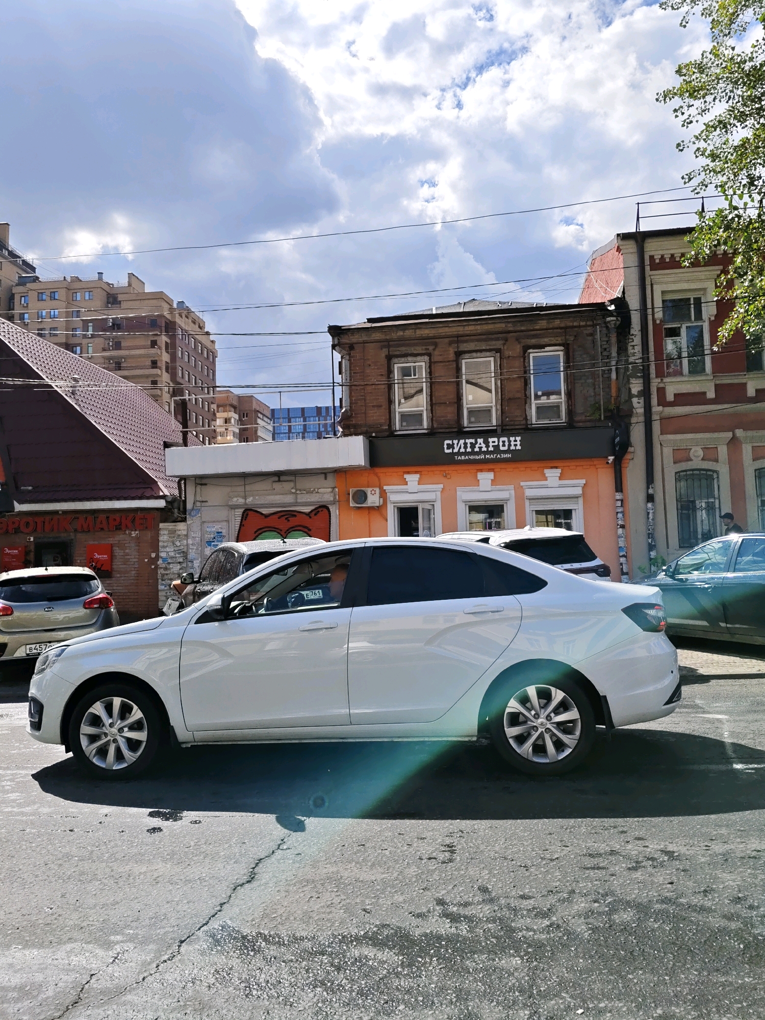 The image depicts a street scene in Rostov-on-Don, Russia. In the foreground, a white sedan is parked, partially obscuring a cracked asphalt road with water puddles and shadows cast by the sun. Behind the sedan, a silver Kia hatchback is visible, with a license plate starting with "B457". To the right, a dark green car is parked.
Further back, a multi-story brick building with a prominent red facade and white trim is visible. Adjacent to it is a two-story wooden building with dark wood siding and several windows. Below the wooden building, an orange-colored structure houses a shop with the Cyrillic text "СИГАРОН" (SIGARON) and "ТАБАЧНЫЙ МАГАЗИН" (TOBACCO SHOP) displayed on a dark awning. The shop has white-trimmed windows and what appears to be an air conditioning unit on its exterior.
To the left of the orange shop, another building with a dark, sloped roof is present, featuring a sign with Cyrillic text including "ЭРОТИКМАРКЕТ" (EROTIC MARKET). This building also has a silver Kia hatchback parked in front of it. In the background, modern multi-story apartment buildings with many windows can be seen. The sky is partly cloudy with a bright sun casting shadows. A person in a black baseball cap is walking on the sidewalk to the right of the orange building.