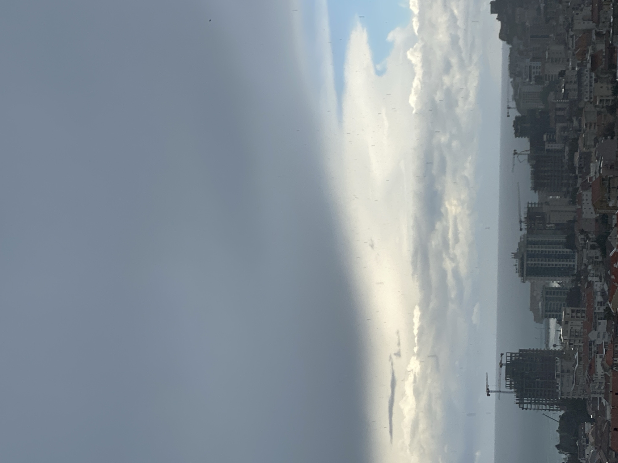 This image provides an elevated, wide-angle view of the city of Budva, Montenegro, under a heavily overcast sky with visible precipitation. The upper half of the frame is dominated by a dark grey cloud layer, from which numerous rain streaks descend, particularly visible against lighter, textured clouds in the middle ground. A small segment of blue sky is discernible in the upper right quadrant.
The urban landscape in the lower half of the image features densely packed buildings. In the immediate foreground, numerous residential structures with reddish-brown tiled roofs are evident. Further back, toward the coastline, several multi-story modern buildings are interspersed with multiple active construction sites. A prominent, tall, unfinished concrete high-rise with a visible tower crane is located in the left-center, adjacent to the sea. Another tower crane is present to the right, overseeing additional construction.
The Adriatic Sea forms the horizon, appearing a muted grey-blue under the cloudy conditions. A harbor or marina with breakwaters and faint outlines of vessels is present in the distance. On the far right, an elevated landmass is topped by dark, irregular structures consistent with ancient fortifications or ruins. No individuals are clearly discernible within the frame.