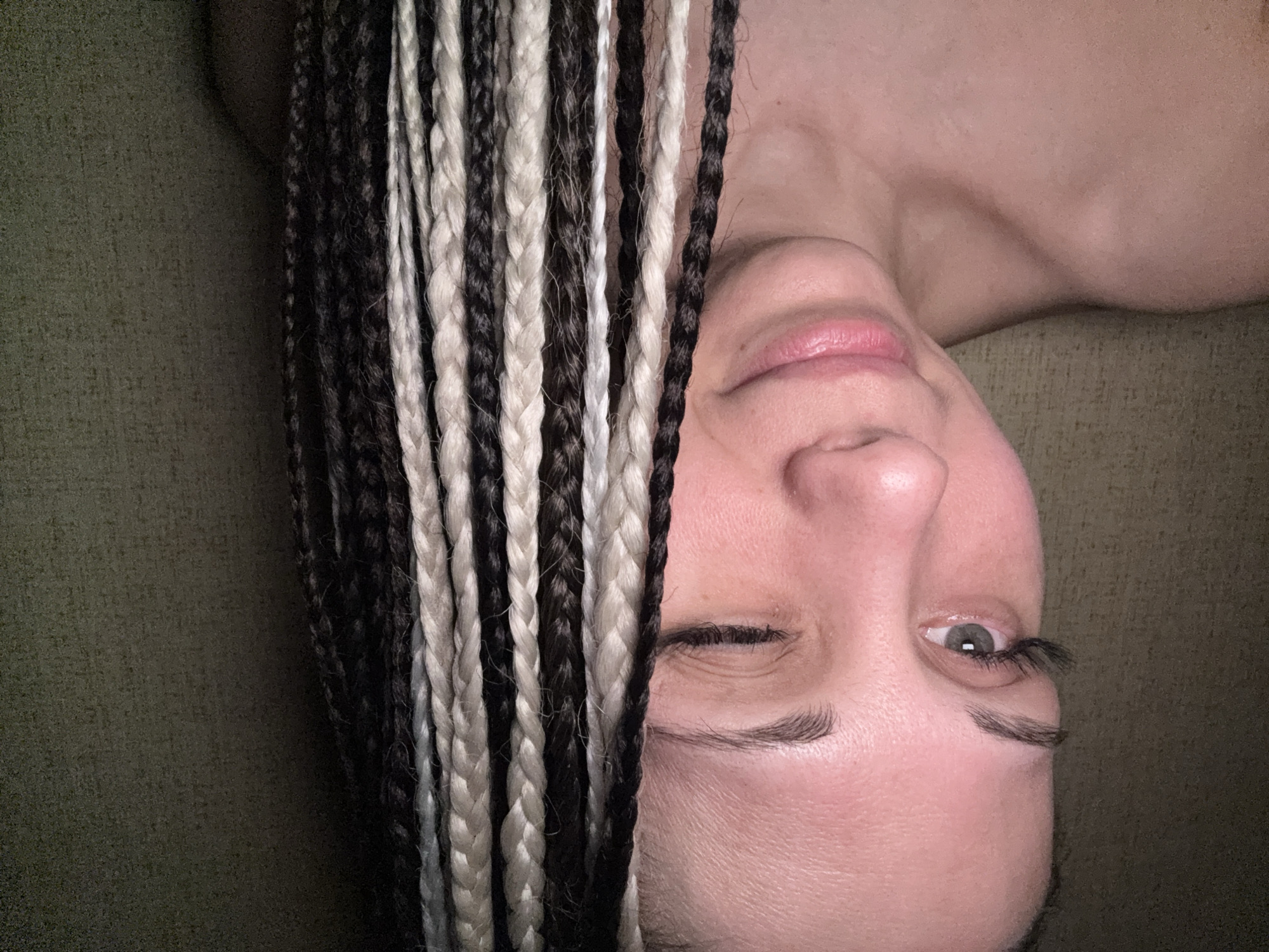 A fair-skinned person is lying on a green surface, possibly fabric. They are facing the camera with their head tilted. The person's hair is styled in dark and light-colored braids that partially cover the left side of their face. They are winking with their left eye. The background is blurred, with only the green surface clearly visible. The location is not identifiable as Rusanovo, Russia based on the image.