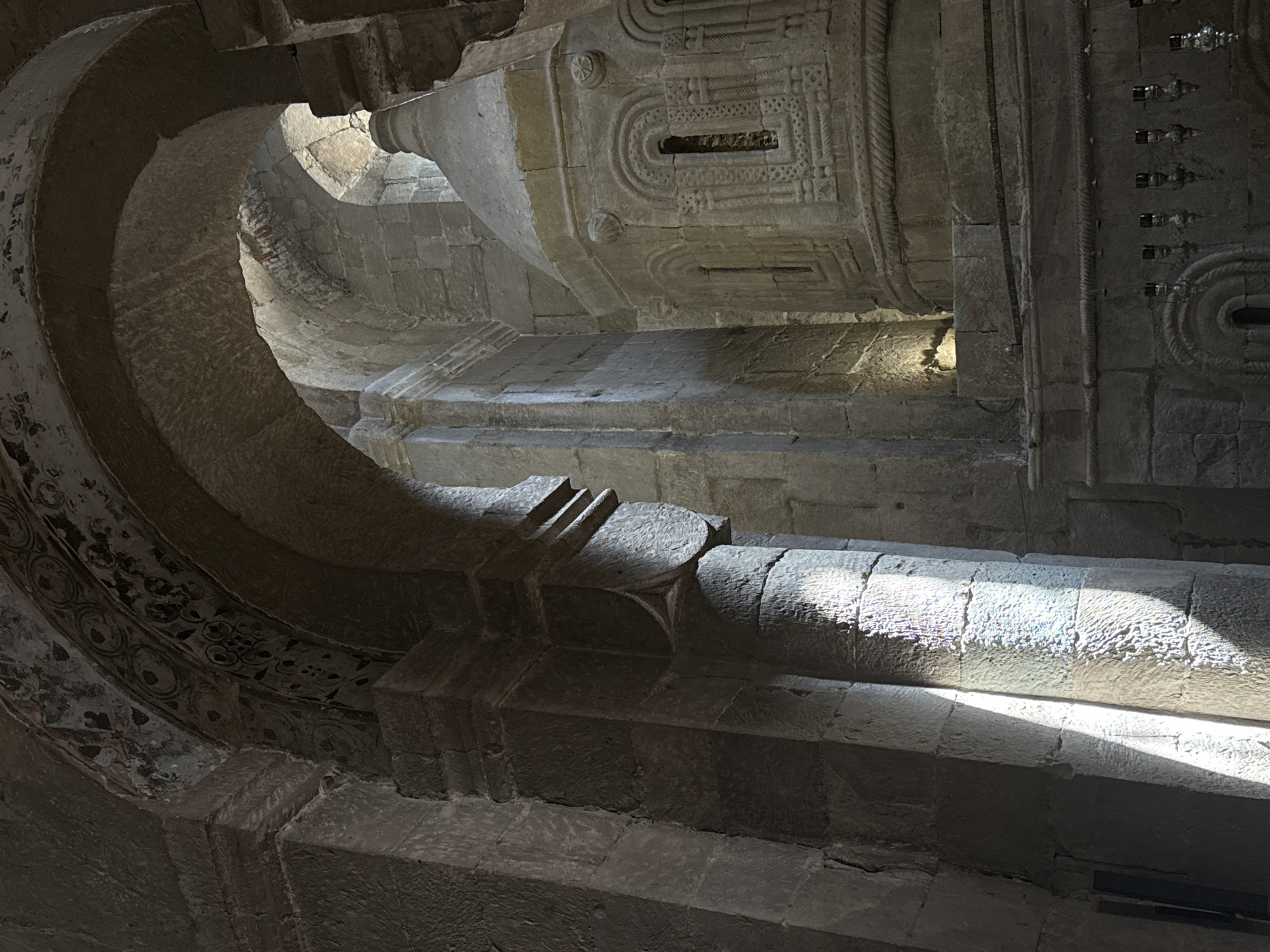 The image shows the interior of a stone church in Mtskheta, Georgia. Sunlight streams through an archway, illuminating a large stone column on the left. The light creates a sharp shadow that falls across the column. In the background, a domed structure with intricate carvings and small windows is visible, also made of stone. Decorative patterns are visible on the archway. The environment is dimly lit, suggesting it is either early morning or late afternoon, or that the church has limited natural light sources. There are no people or animals in the frame. The scene depicts a historical and architectural interior.