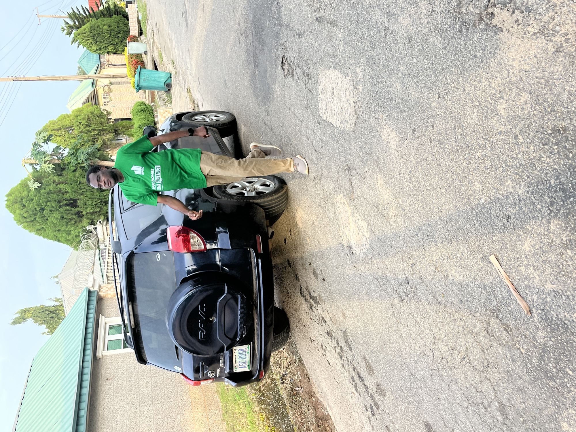 A man in a green t-shirt and tan pants walks on a paved road next to a black Toyota RAV4 SUV. The vehicle's rear is visible, including a spare tire cover and a license plate that reads "ABC-886R". The man is walking away from the camera, his head turned to his left. The scene is set in a residential area with houses and trees. There are two green trash cans visible in the background. The road appears to be in a state of disrepair with patches of exposed aggregate. The sky is overcast. This scene is located in Abuja, Nigeria.