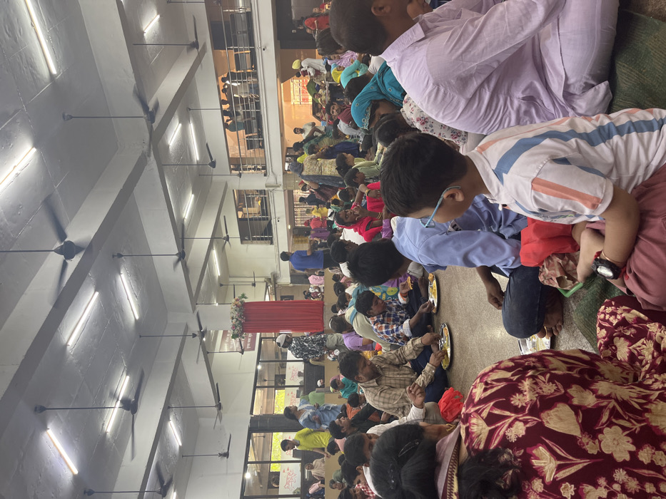 The image shows a large gathering of people in a hall with a high ceiling.  The hall appears to be a public space for a community gathering in Panchkula, India. The location is on the ground floor of a multi-story building at the intersection of Sector 12A, 12, and 11 in Panchkula, India (latitude: 30.723, longitude: 76.862). Many ceiling fans hang from the ceiling, keeping the air moving in the space. The people are sitting on the floor in rows, eating from plates, and interacting with each other. They are dressed in a variety of clothing. There is a red curtain and a floral arrangement in the background. The scene is a public gathering of people, possibly for a celebration or a meal.