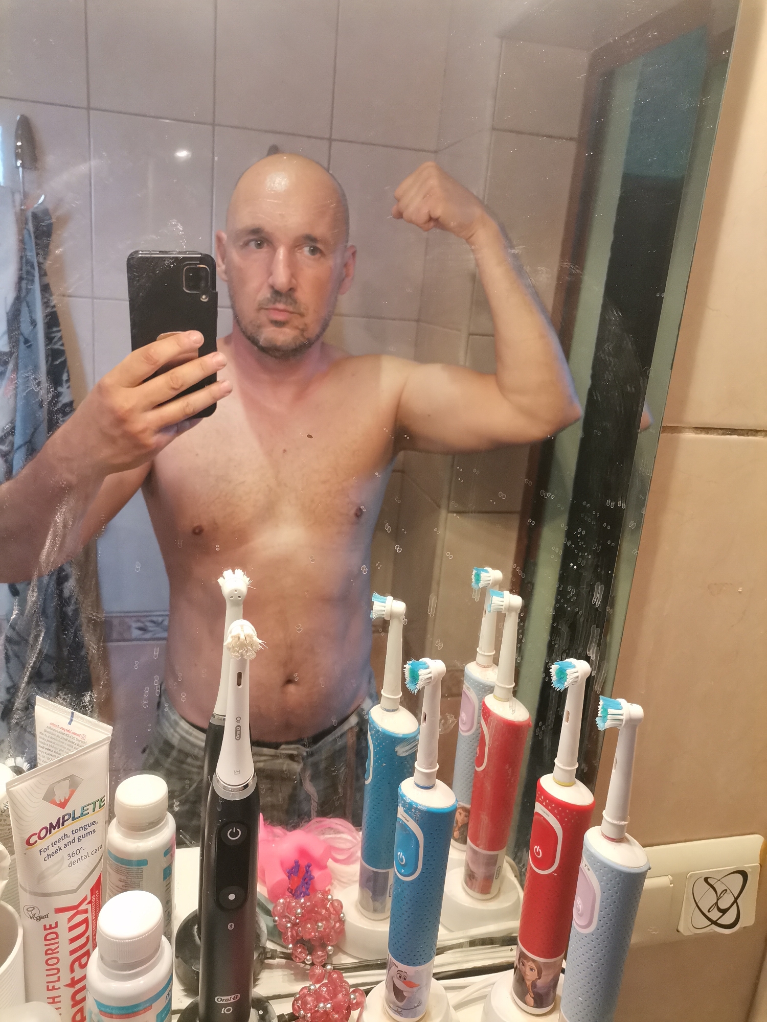 The image depicts a shirtless, bald man taking a selfie in a bathroom mirror. He is flexing his right arm. The bathroom has tiled walls, and the mirror has water spots. In front of the mirror, on the counter, are several electric toothbrushes, toothpaste tubes, and other toiletries.