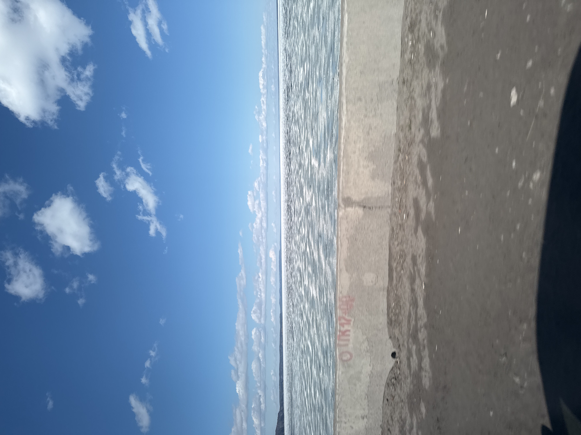 The image depicts a waterfront scene in Chaikovsky, Russia. The scene shows a body of water, possibly a river or reservoir, with a concrete embankment in the foreground. The sky above is bright blue with scattered white clouds. Graffiti is visible on the concrete embankment. A dark, indistinct object is present in the lower right corner of the frame.