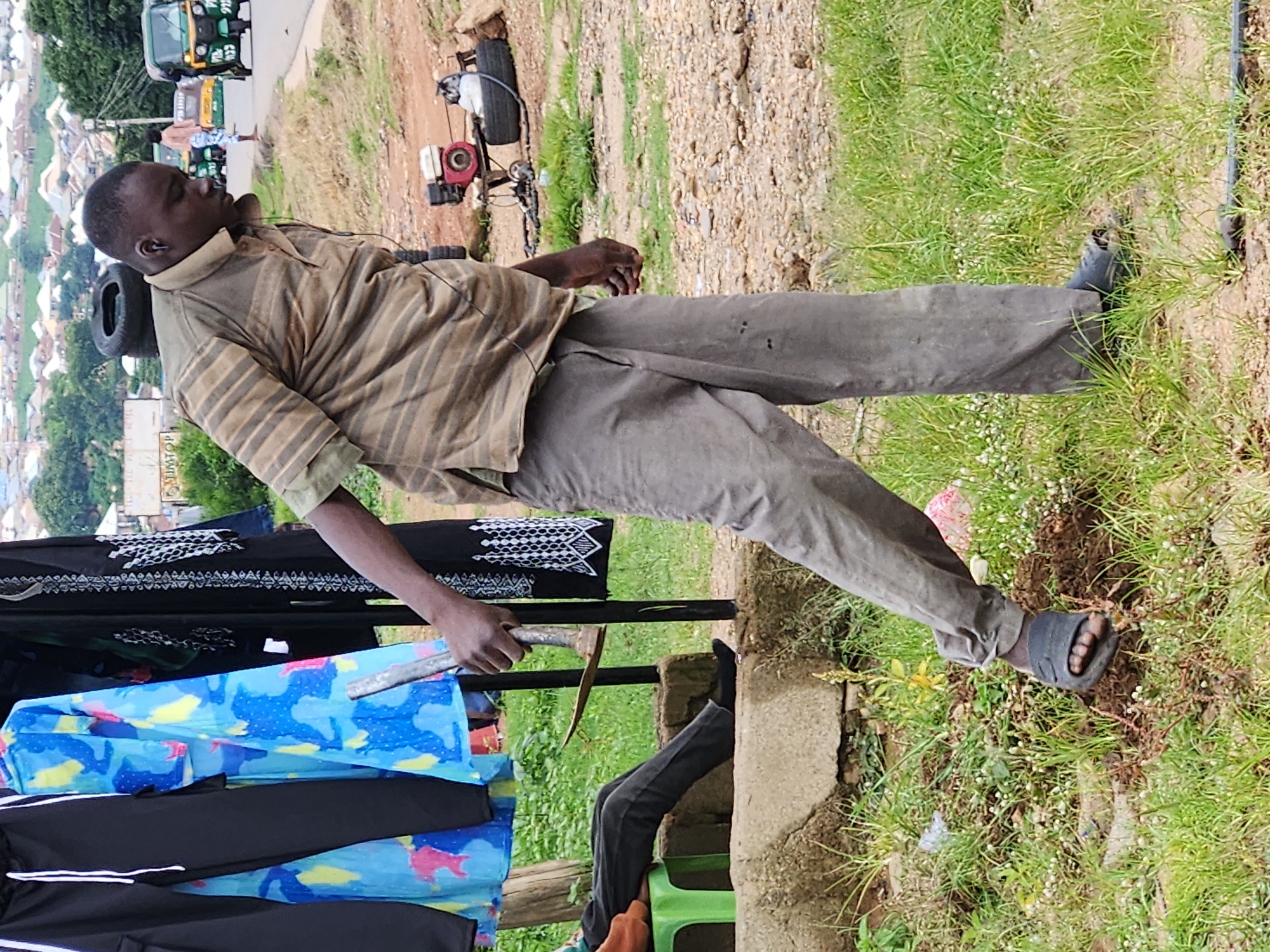 The image captures a scene on Rukuba Road, Nigeria, with a man in a striped shirt and grey pants holding a tool that appears to be a pickaxe. He is standing on a dirt path, with green grass and weeds growing around him. In the background, there are several vehicles, including what looks like a tuk-tuk, and a distant view of buildings. To the left of the man, there are clothes hanging on a rack, with colorful patterned fabric visible. A person is sitting on a green stool to the far left, wearing dark pants. There is also a small engine and tires stacked on top of each other in the middle ground. The overall environment suggests an outdoor, possibly semi-urban setting.