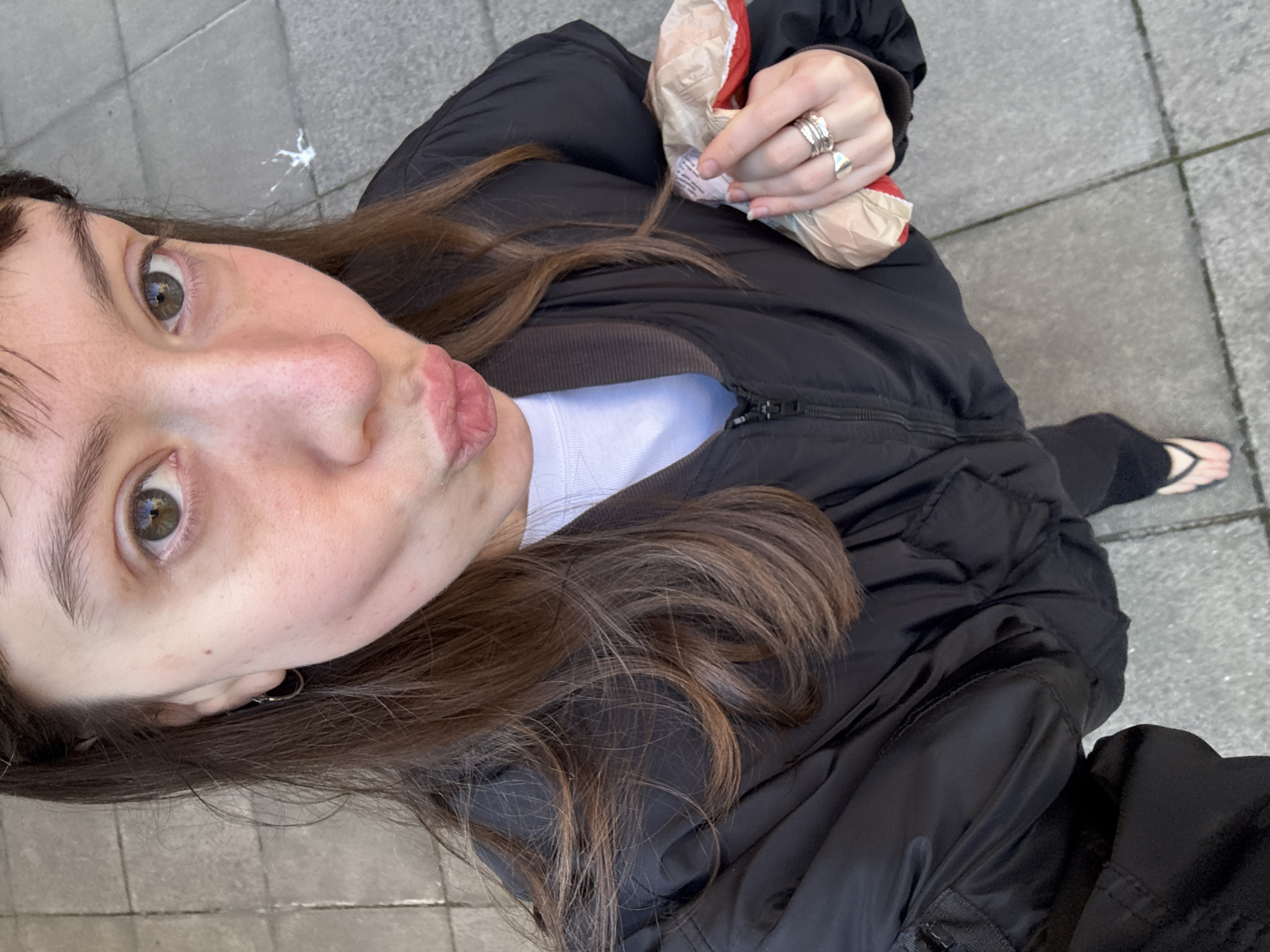 A woman stands outdoors on a gray paved surface, likely a sidewalk, holding a crumpled paper bag in her left hand. She wears a black puffer jacket, a white t-shirt, and black pants or leggings. Her long brown hair is partially obscuring her jacket. On her left hand, she has several rings. She is looking directly into the camera with her lips pursed. In the background, the pavement suggests an urban setting. The presence of a splattered white substance on the ground near the top left corner suggests it is a street in London. A small portion of her right foot and flip flop is visible. The photo is taken from an overhead, selfie-style perspective.