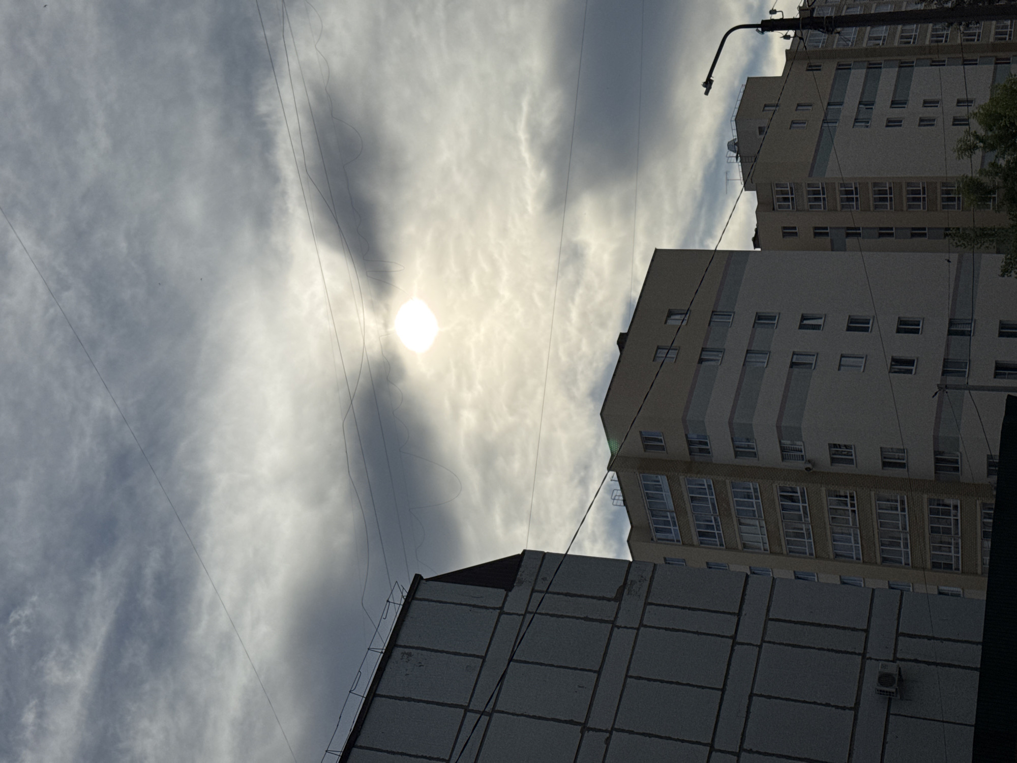 The image shows a view of apartment buildings under a cloudy sky with the sun partially obscured by clouds. In the foreground on the left is the corner of a light-colored building with visible horizontal and vertical lines suggesting construction blocks. A black cable runs diagonally across the frame from the top left to the bottom right. To the right of this building, two taller, multi-story apartment buildings are visible. The building in the middle ground has a light yellow facade with horizontal bands of gray and white, and numerous windows. The building further to the right also features a similar design with a light facade and banded detailing. A street lamp with a dark pole and light fixture stands to the right of this second apartment building. The sky is filled with a mix of dark and light gray clouds, with the sun appearing as a bright, hazy orb in the upper center of the frame. More black cables are strung across the sky. The scene depicts a typical urban residential environment in Chaikovsky, Russia.