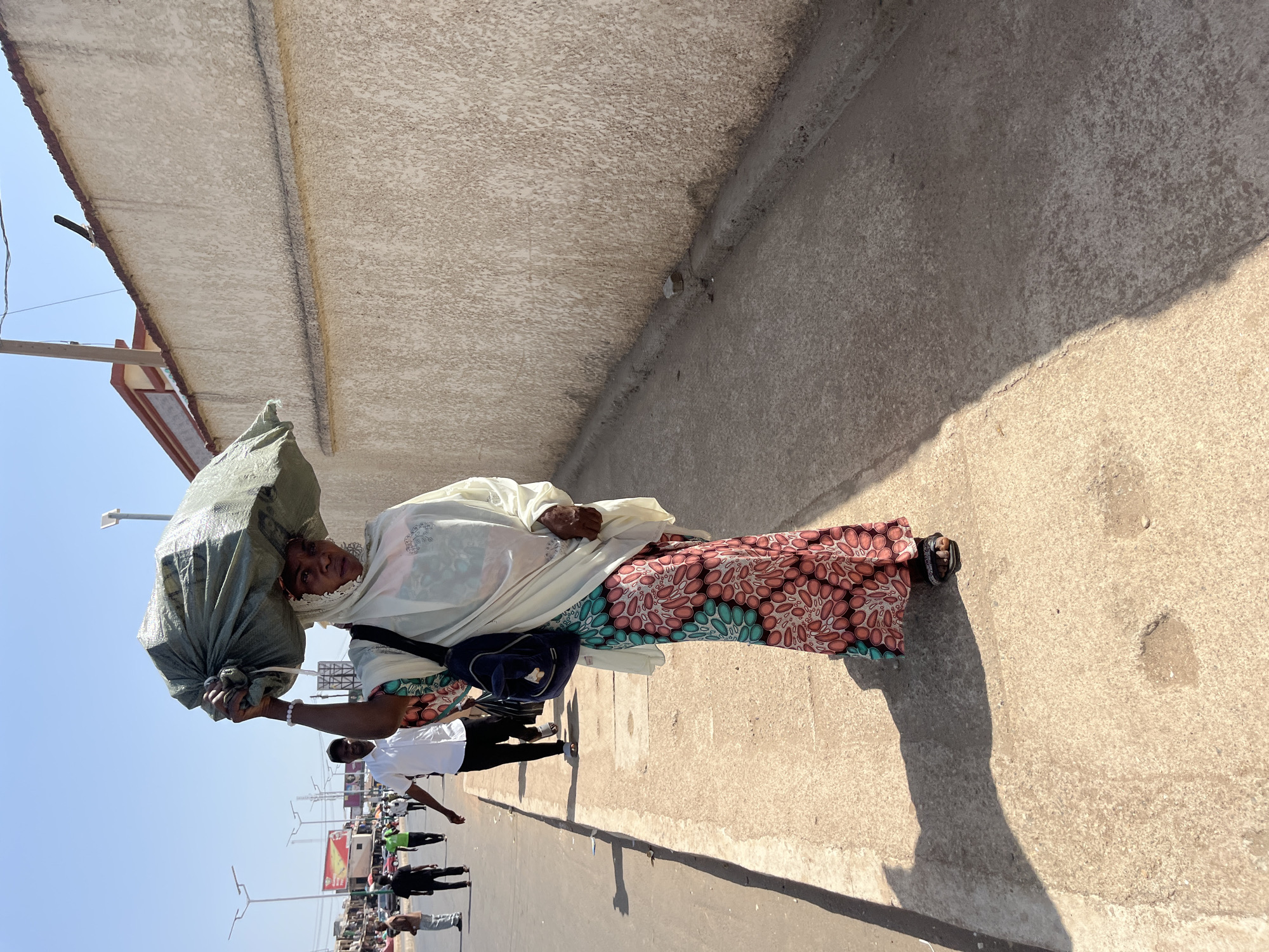 An African woman carries a large green sack on her head while walking on a concrete sidewalk next to a street in Jos, Nigeria. She is wearing a white headscarf and a patterned dress. Several people are walking in the opposite direction on the street. A building is visible on the left side of the frame. Streetlights and commercial signs are also visible in the background.