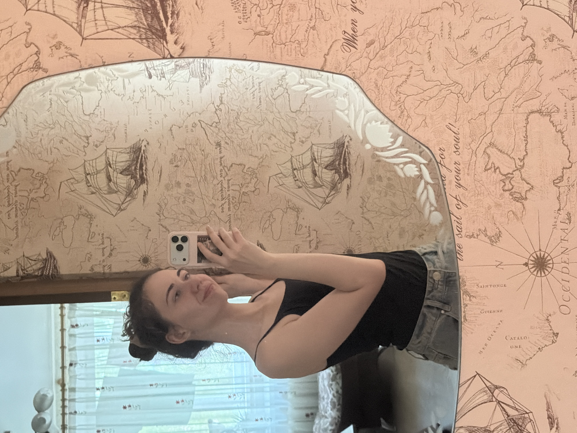 A young woman with dark hair pulled into a bun takes a mirror selfie. She is wearing a black tank top and light-wash denim shorts. The background shows a wall with a patterned wallpaper featuring old maps and sailing ships. A window with light-colored curtains is visible in the reflection to her left. The image is a portrait of the woman, taken in an indoor setting, likely a bedroom, in Zavodoukovsk, Russia.