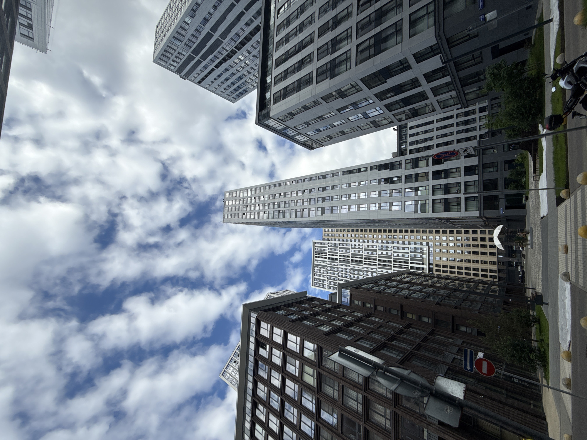 The image shows a view looking upward at several modern buildings against a cloudy sky. The buildings are of varying heights and designs, some with dark facades and others with light gray or white facades. Many windows are visible. On the right side of the frame, a sidewalk with a street lamp is seen, and a glimpse of a motorcycle is visible. A no entry road sign is visible near the bottom of the frame. Based on the architectural style and known landmarks, the image is located in the Moscow International Business Center (Moscow City), Russia.