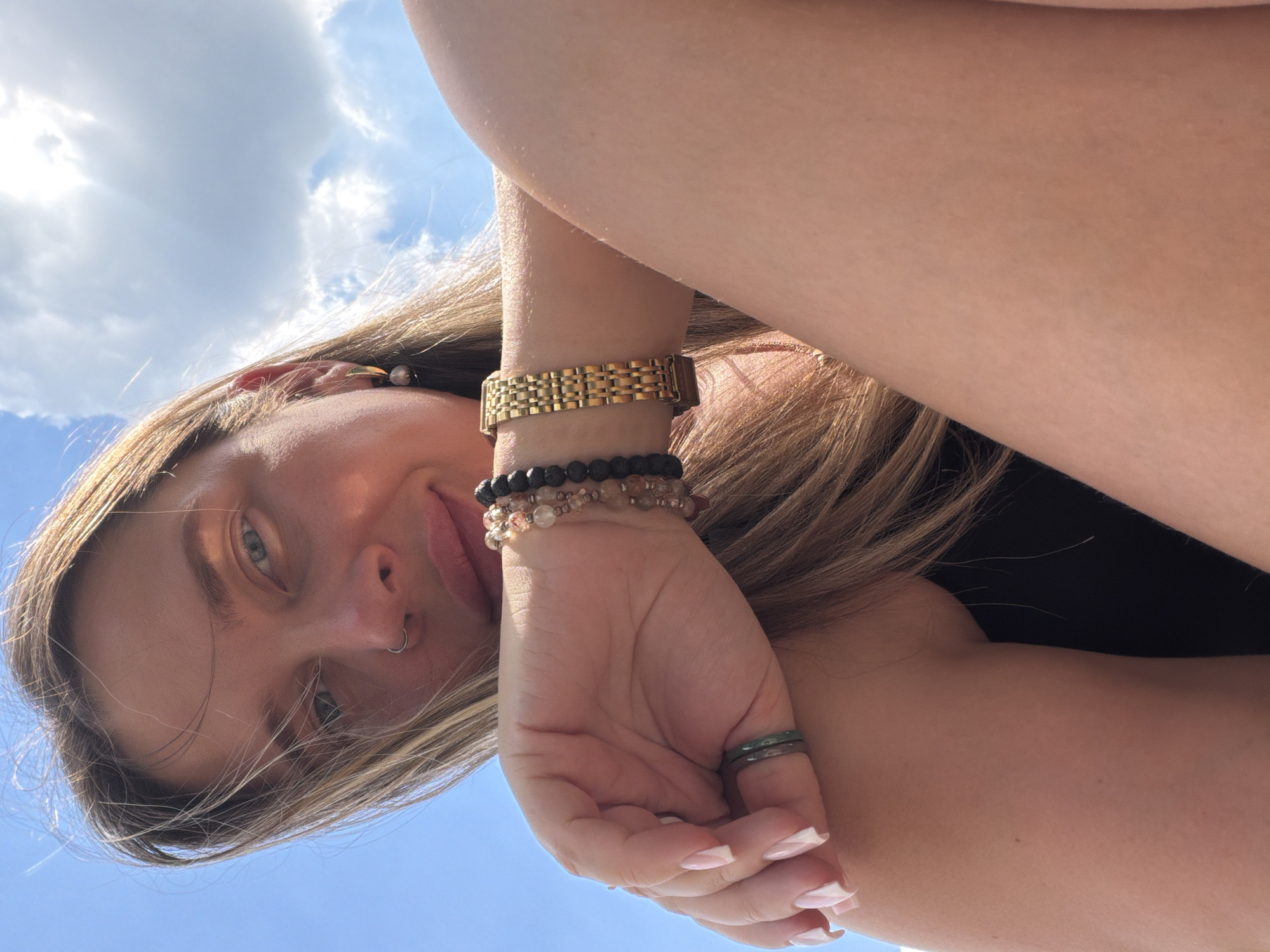 A close-up, low-angle, outdoor selfie of a young woman with blonde hair, wearing a black top, gold watch, and bracelets, as well as rings on her fingers, is captured against a bright blue sky with clouds. The woman is looking directly at the camera with a slight smile. The scene does not provide any specific details to indicate a location in Moscow, Russia.