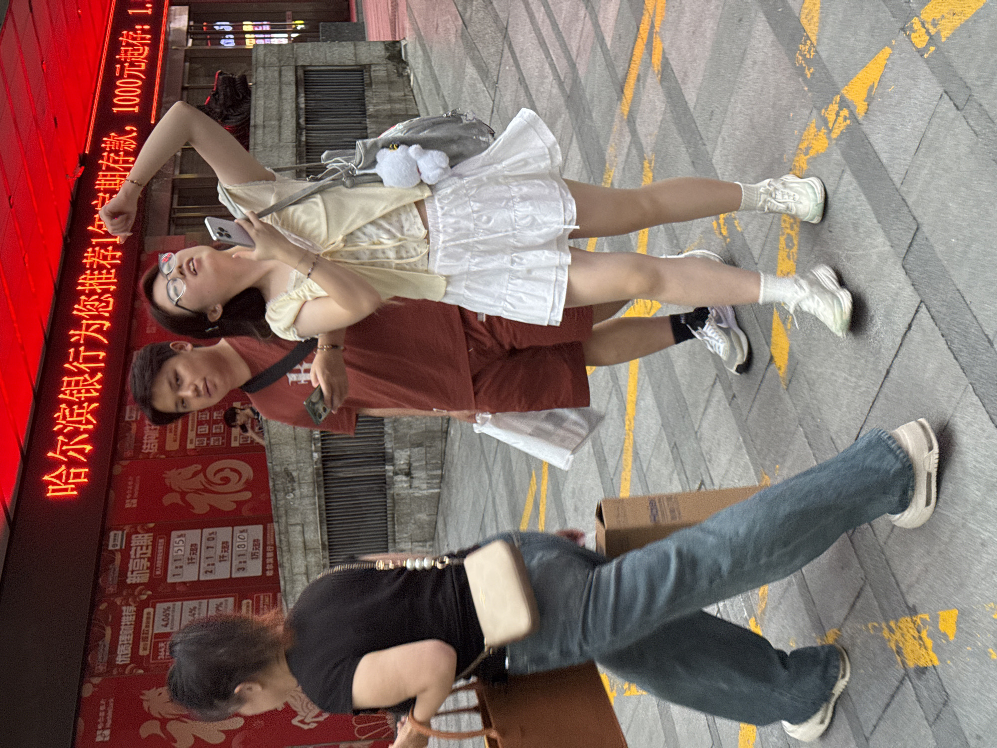 In Yuzhong District, China, a group of people walk on a city street. A woman in the foreground, facing left, wears a black t-shirt and blue jeans, carrying a brown tote bag in her left hand and a small beige crossbody bag. Behind her, a man in a reddish-brown t-shirt and shorts holds a smartphone. To his right, a woman in a light yellow top and white skirt looks up, holding a smartphone in her right hand and a grey bag with a white teddy bear charm on her left shoulder. The background features a red neon sign with Chinese characters advertising "Harbin Bank" and financial services. The street is paved with grey tiles marked with yellow lines, and a cardboard box sits on the ground behind the woman in blue jeans.