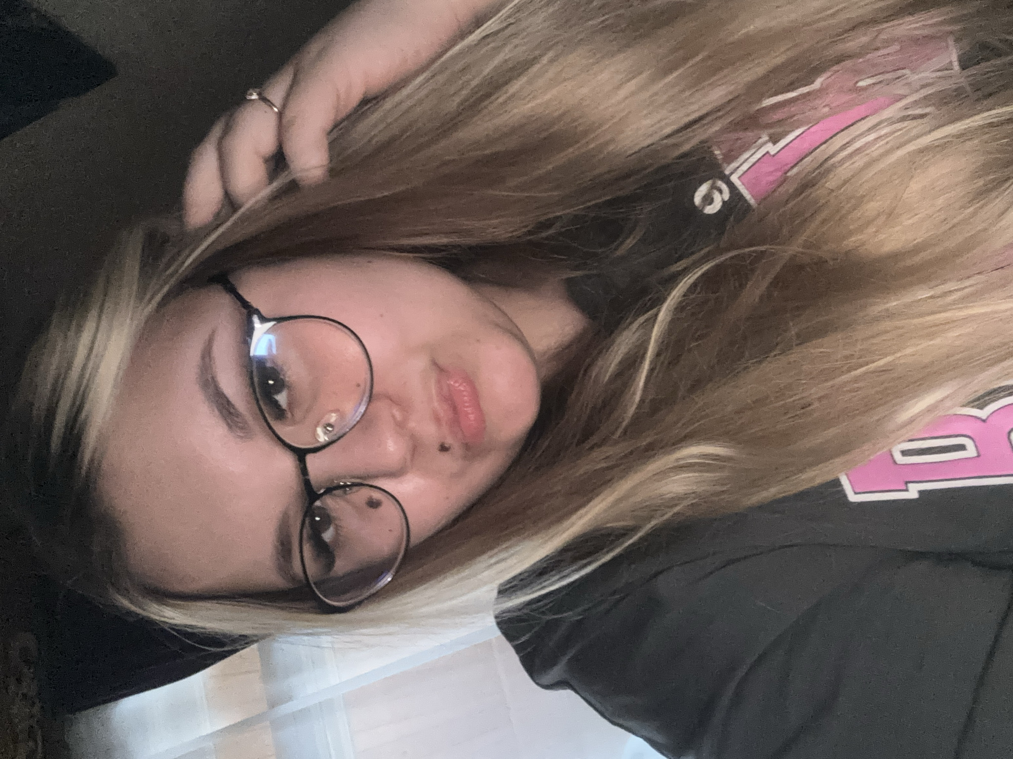 A young woman with blonde hair and glasses is posing for a selfie. She is wearing a dark grey hoodie with pink lettering on it. Her hair is long and wavy, and she is touching it with her right hand. She has two moles on her face, one on her cheek and one near her lip. The background appears to be indoors, with sheer curtains visible in the upper left corner. There are no specific indicators in the image that would suggest a location in the city of Bolshoy Bukor, Russia.