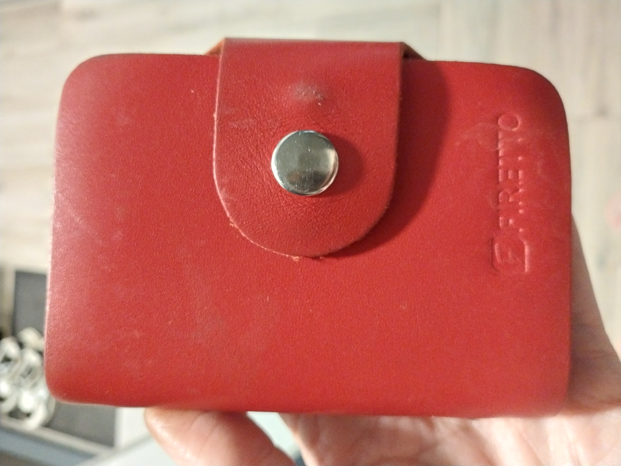 The image shows a red, textured cardholder held in a person's hand. The cardholder has a snap closure with a silver button. The brand name "FIRETTO" is embossed on the bottom right corner of the cardholder. The background is blurry but appears to be an indoor setting with a light-colored floor and a white object in the upper left corner. The image was taken in Dnipro, Ukraine.