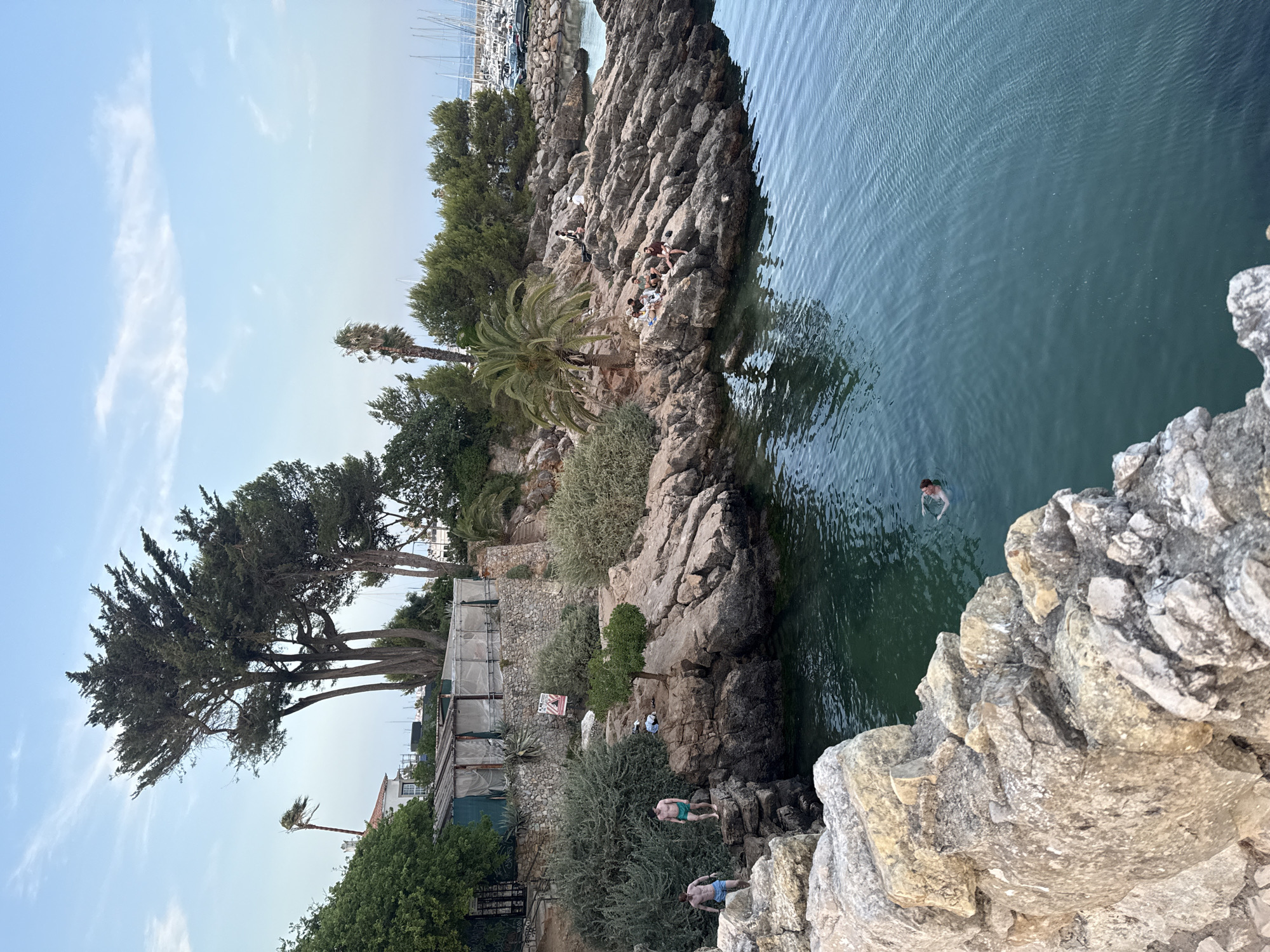 The image depicts a scenic coastal area in Cascais, Portugal. In the foreground, large, rough-hewn rocks line the edge of the clear, turquoise water. Two young men in swim trunks stand on the rocks on the left side of the image, seemingly preparing to enter the water. Further along the rocky coastline, a group of people are seated and lounging on the sun-drenched rocks, some with their legs dangling towards the sea. A palm tree provides shade for some of the individuals.
In the background, a building with a rustic, stone facade and a covered outdoor area with white fabric awnings sits atop a rise, surrounded by lush green vegetation. A tall, windswept tree stands prominently near the building, its branches reaching towards the sky. Beyond the rocky shore, a marina filled with numerous sailboats and yachts is visible under a partially cloudy, pale blue sky. The overall scene suggests a relaxed, warm-weather atmosphere at a popular seaside location.
