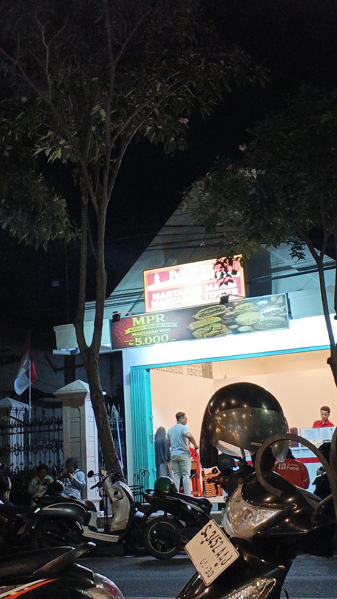 A nighttime outdoor scene in Bojonegoro, Indonesia, depicts a food stall with bright lighting, attracting customers. The stall features a prominent sign that reads "MPR MARTABAK" and advertises "MARTABAK TEFLON" and "MARTABAK MINI" with a price of "Rp 5.000". Images of martabak are displayed on the sign. Several motorcycles are parked in front of the stall, some with helmets on them. A few individuals are visible inside and outside the stall, with one person in a light blue shirt standing at the counter and another in a red shirt potentially working. The environment is dimly lit by the stall's lights, with trees casting shadows and a dark sky above. A small Indonesian flag is visible on the left side of the image. The overall impression is a bustling local eatery at night.