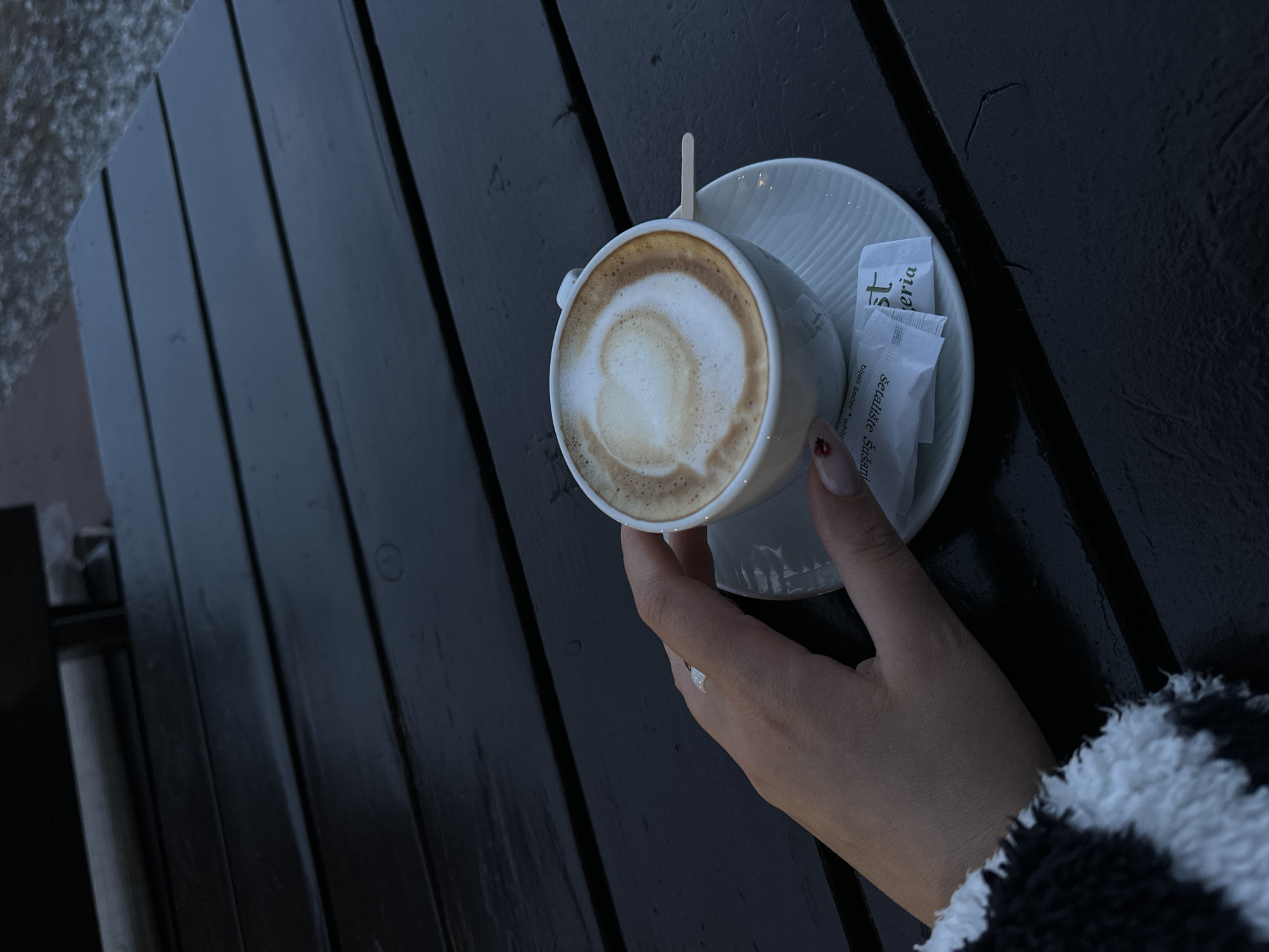 An overhead shot captures a person's hand holding a white ceramic coffee cup and saucer on a dark wooden slatted table, likely outdoors. The cup contains a beverage, presumably coffee, with latte art in the form of a heart or abstract swirl on its frothy surface. A small wooden stirrer stick is placed in the cup.
Next to the cup, on the saucer, are two white sugar packets. The visible text on one packet reads "Šetalište Sušanj," and below that, "bijeli šećer * white." The other packet has "st eria" partially visible.
The hand, adorned with a simple ring on the ring finger, has neatly manicured nails, with one nail (pinky or ring finger) showing a small red dot or detail. The person is wearing a black and white fuzzy or fleece sleeve, suggesting cooler weather.
The wooden table appears dark and slightly damp or aged, with subtle textures and reflections. To the left, a blurry, lighter, granular surface is visible, possibly concrete or a path, hinting at an outdoor setting. The lighting is subdued, suggesting an overcast day or late afternoon. The overall scene conveys a quiet moment of enjoying a warm drink. The location is Bar, Montenegro.