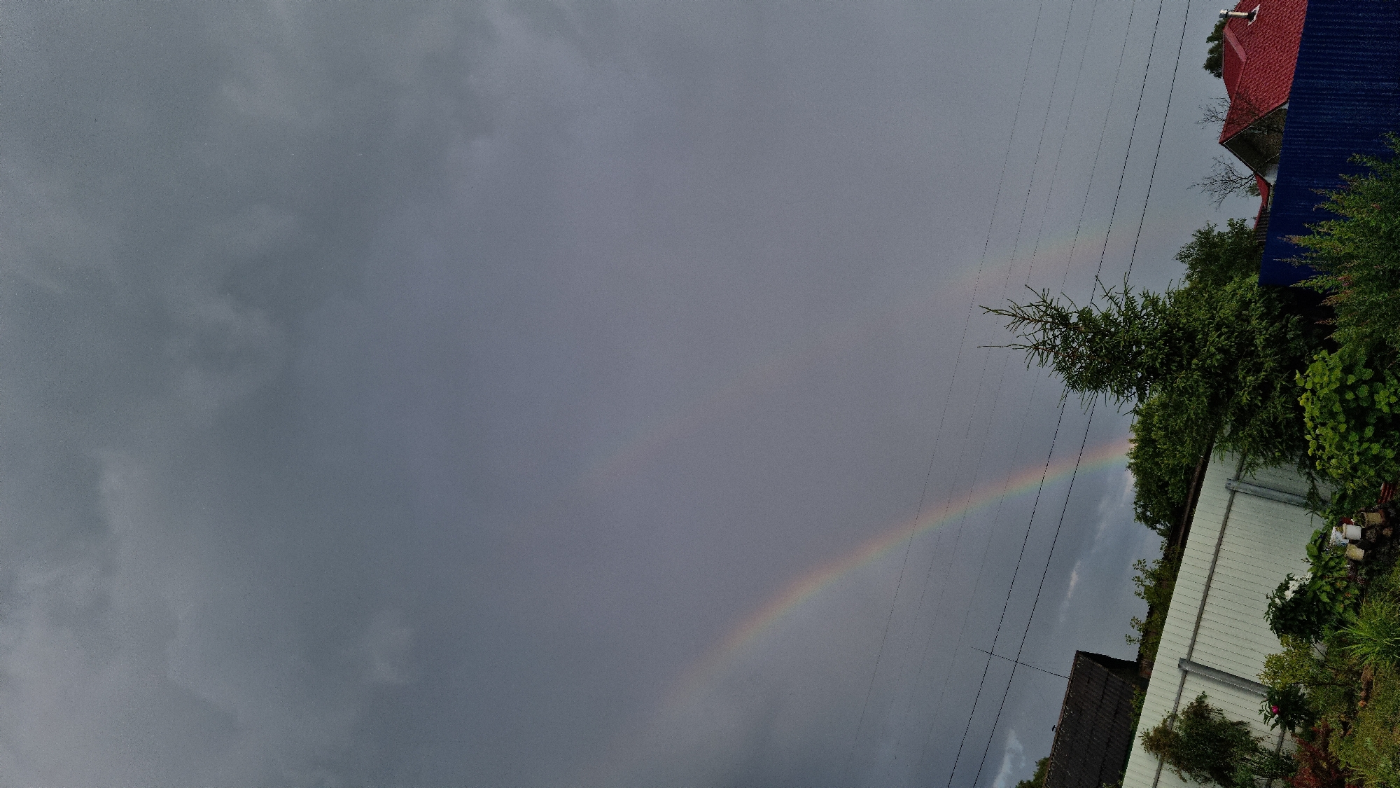 This is a photograph of a sky with a rainbow, taken in Vysokoe, Russia. The sky is overcast with dark, gray clouds, suggesting it might have recently rained or is about to. A faint rainbow arc is visible in the middle of the sky, spanning from the left side of the frame towards the right.
In the foreground, on the right side of the image, are parts of buildings and lush green foliage. There is a white structure, possibly a shed or a garage, with a ridged white siding. Above this, a house with a bright red roof and a dark blue, corrugated metal roof extension is visible. Several trees and bushes with dense green leaves are also present, some appearing quite large. Power lines are stretched across the upper right portion of the frame, running diagonally downwards.
The scene appears to be an outdoor residential area, possibly a backyard or a street view. The lighting is dim due to the heavy cloud cover, but the rainbow offers a touch of color and contrast to the otherwise muted tones of the sky and landscape. There are no people visible in the image. The time of day is likely late afternoon or early evening, given the cloud cover and the subdued light. The overall mood of the image is somewhat somber due to the weather, but the presence of the rainbow adds a hopeful or serene element. No text is visible in the image.