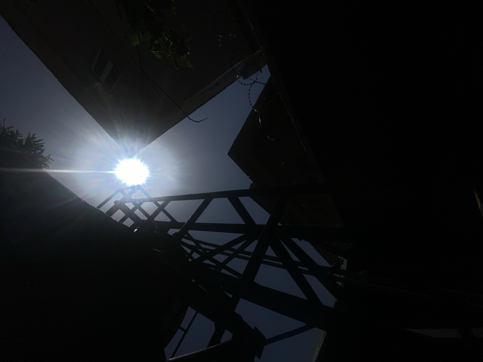 The image is a low-angle shot of a courtyard in Barden Yana, Nigeria, taken on a sunny day. The sun is visible in the top left corner, and its rays shine through the gaps between the buildings and a metal framework. The buildings are silhouetted, creating strong contrasts between light and shadow. Wires and vines are visible near the buildings. The metal framework is in the center of the image and reflects on a surface below. The right side of the image is dominated by a dark, shadowy area.