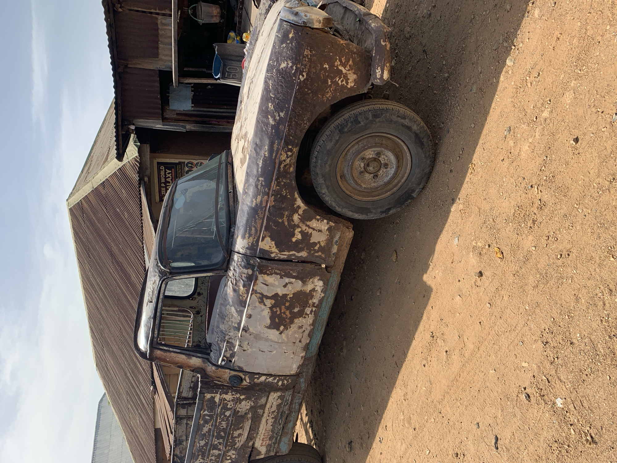 The image shows a severely rusted and dilapidated light truck parked on a dirt surface in Rafin Par, Nigeria. The vehicle's paint is heavily peeling, revealing rust on most of the body panels. One tire is visible, appearing worn. The truck is situated beside a building with a corrugated metal roof and weathered wooden walls. A sign reading "Zee P World Galaxy" is visible on the building. The scene conveys a sense of neglect and disrepair.