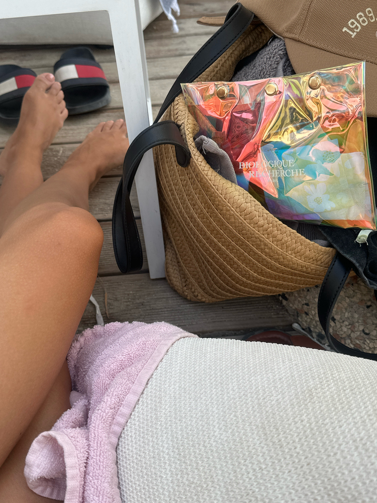 The image captures a close-up view of a person's legs and feet, resting on a wooden deck. To the left, a pair of black slides with red and white accents are visible. A straw bag, containing a transparent, iridescent pouch with the text "BIOLOGIQUE RECHERCHE PARIS" printed on it, is positioned to the right. A tan baseball cap with the year "1989" embroidered in white is also peeking out of the bag. A pink towel is draped over the person's legs. The overall scene suggests a relaxed setting, possibly outdoors. The location is identified as Pallini Municipal Unit, Greece.