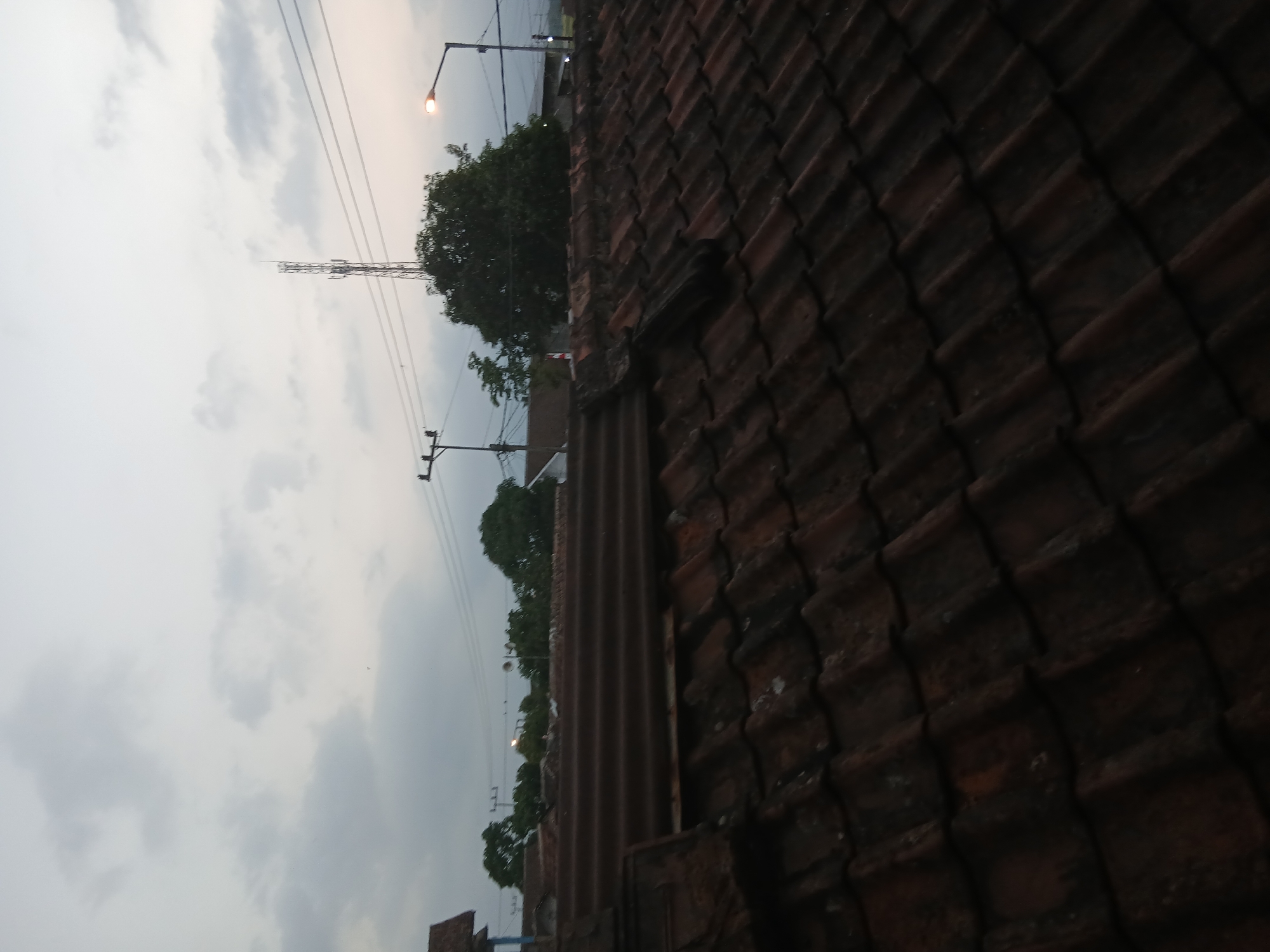 The image captures a rooftop view in Tenggarang, Indonesia, during what appears to be twilight or overcast conditions. The foreground is dominated by a weathered, reddish-brown tiled roof, with individual tiles showing signs of age and some light discoloration. In the mid-ground, a slightly different type of corrugated roofing material is visible, also showing signs of wear. Beyond the rooftops, a cluster of trees with dense green foliage stands out. In the distance, a tall communication tower is prominent against the sky, with power lines extending across the frame. A street lamp with an illuminated bulb is also visible on the right side of the image. The sky is a muted gray with scattered clouds, suggesting a lack of direct sunlight. No people or animals are discernible in the scene.