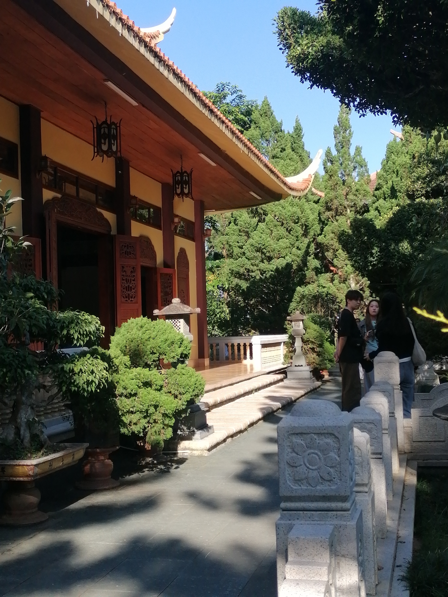 The image depicts a Buddhist temple with traditional Vietnamese architecture, featuring a curved, orange-tiled roof with decorative eaves. The temple walls are painted a pale yellow, with dark wooden accents and ornate doorways. Two dark, hexagonal lanterns hang from the eaves. In the foreground, a pathway is lined with decorative stone balustrades featuring floral carvings. To the left, there are meticulously pruned bonsai trees in decorative planters.
In the mid-ground, three individuals are standing and conversing near a stone lantern. A young man with dark hair, wearing a black t-shirt and brown pants, is facing left. Beside him, a young woman with long dark hair, wearing a light blue top, is looking towards the man. To her right, another individual with long dark hair, wearing a dark top and blue jeans, is also looking towards the man. Behind them, lush green trees and foliage surround the temple, suggesting a garden setting.
The overall scene appears to be tranquil and serene, typical of a Buddhist temple or pagoda. The presence of people suggests it is a place accessible to visitors, possibly a tourist attraction or a place of worship. The bright sunlight and clear blue sky indicate a pleasant day. This scene is consistent with the architecture and landscaping found at the Truc Lam Zen Monastery in Da Lat, Vietnam, a prominent Buddhist site known for its serene environment and beautiful gardens.