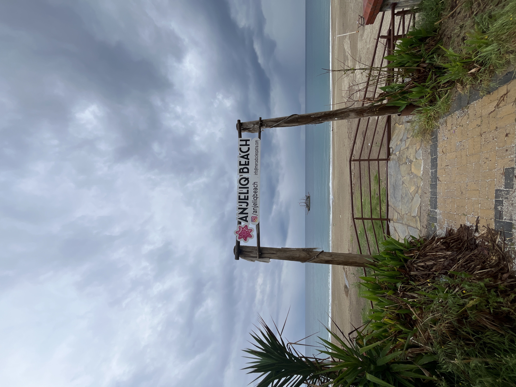 The image shows a beach scene, possibly taken in Alanya, Turkey, as indicated by the signage that reads "ANJELIQ BEACH" with an Instagram handle and a website related to mercado cleopatra. A weathered, wooden signpost marks the entrance to the beach, which leads through a metal gate and a short pathway constructed of brick. The beach itself is sandy, and the sea is calm and grayish. A distant ship is barely visible. The sky is overcast with thick, grey clouds. Vegetation including shrubs and palm-like plants are seen on both sides of the walkway. The photograph is taken from a slightly low angle.