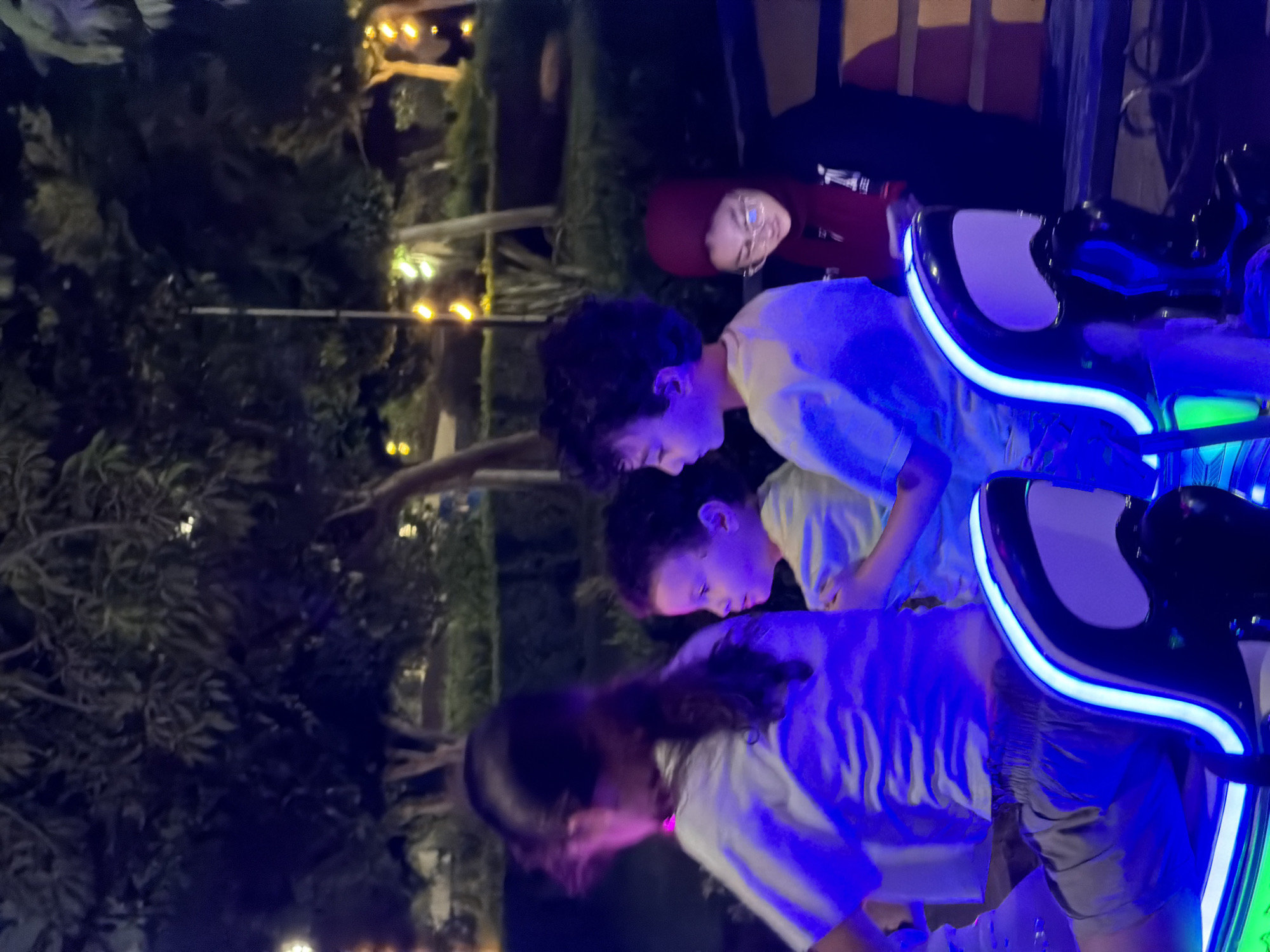 In an outdoor setting at night, possibly in the city of 6th of October, Egypt, four individuals are seated in what appears to be a ride or attraction. The scene is illuminated by blue and green neon lights, creating a vibrant and somewhat surreal atmosphere.
In the foreground, a person with dark hair pulled back is seated on the left, facing away from the camera. They are wearing a white t-shirt and shorts. To their right, a young boy with curly dark hair is seated, looking to the left. He is wearing a light-colored t-shirt. Next to him, another young boy with lighter curly hair is seated, looking to the right. He is also wearing a light-colored t-shirt. On the far right, a person wearing a red hijab and glasses is seated, looking down at something in their hands. They are wearing a black jacket.
The individuals are seated in what looks like custom seats with illuminated edges, suggesting a modern amusement or entertainment experience. In the background, trees are visible, with some illuminated decorative lights in the distance, creating a festive ambiance. The ground is dark, indicating it is nighttime. The overall impression is of a family enjoying an evening out at an entertainment venue.