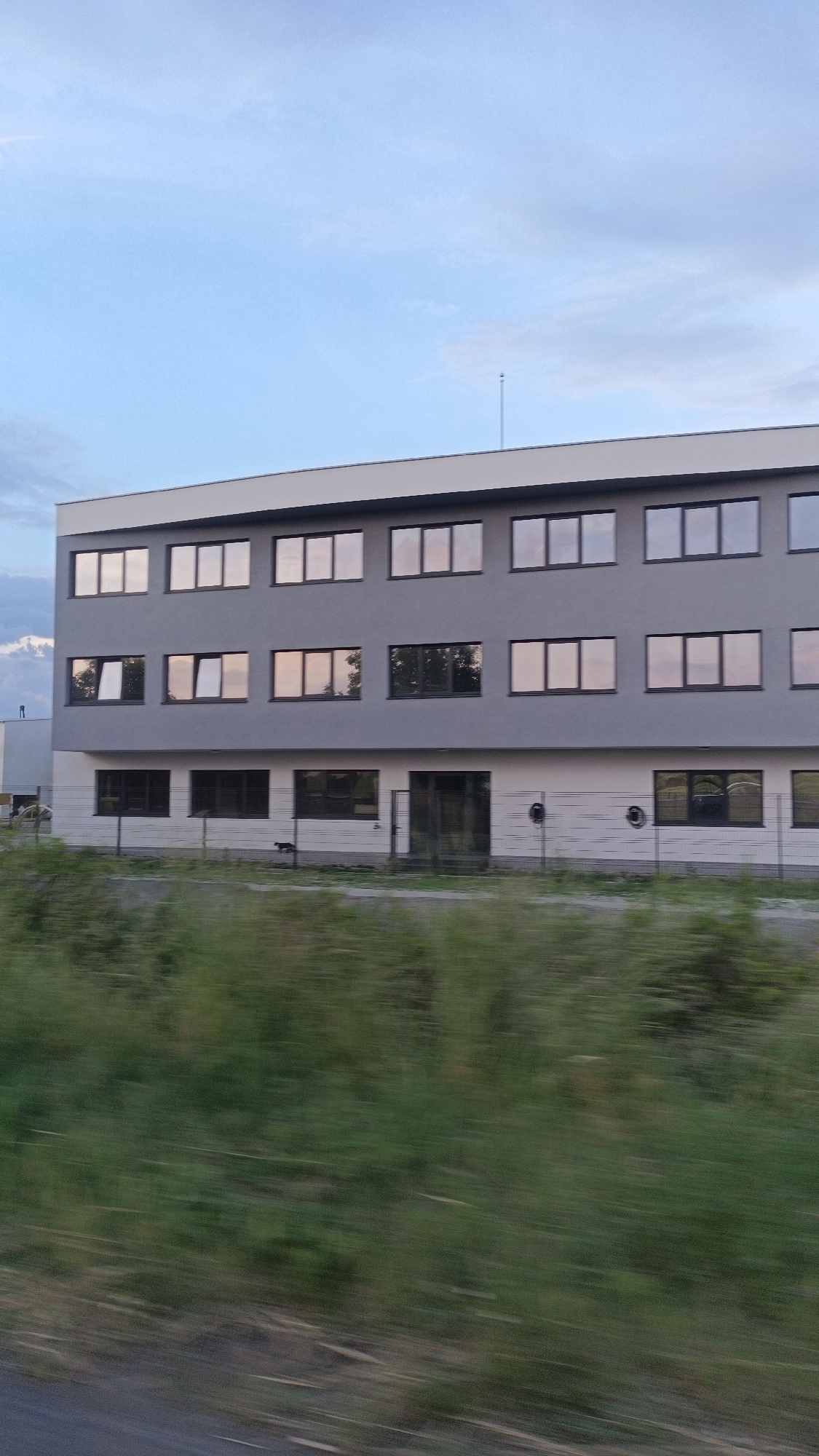The image shows a three-story building in Burgas, Bulgaria. The building has a grey exterior with white trim. There are rectangular windows on the second and third floors. The bottom floor has a mix of windows and a doorway. A chain-link fence runs in front of the building, separating it from a field of green grass and vegetation. The sky is partly cloudy. The image appears to have been taken from a moving vehicle.