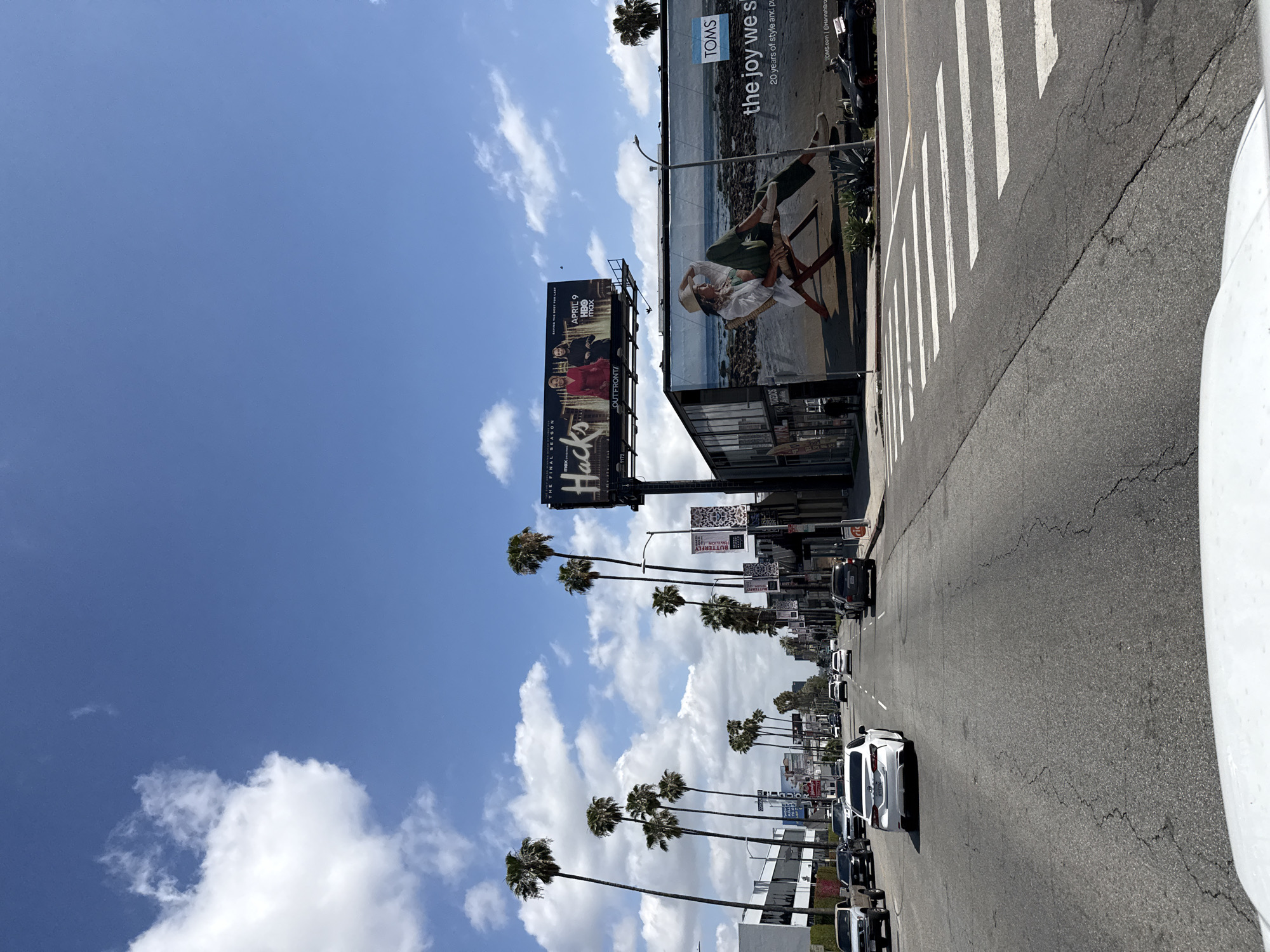 The image shows a street scene in Los Angeles, California. Palm trees line the street, with billboards displaying advertisements for "Hacks" and "TOMS" above. The street is filled with cars, and a crosswalk is visible. The sky is blue with scattered clouds.